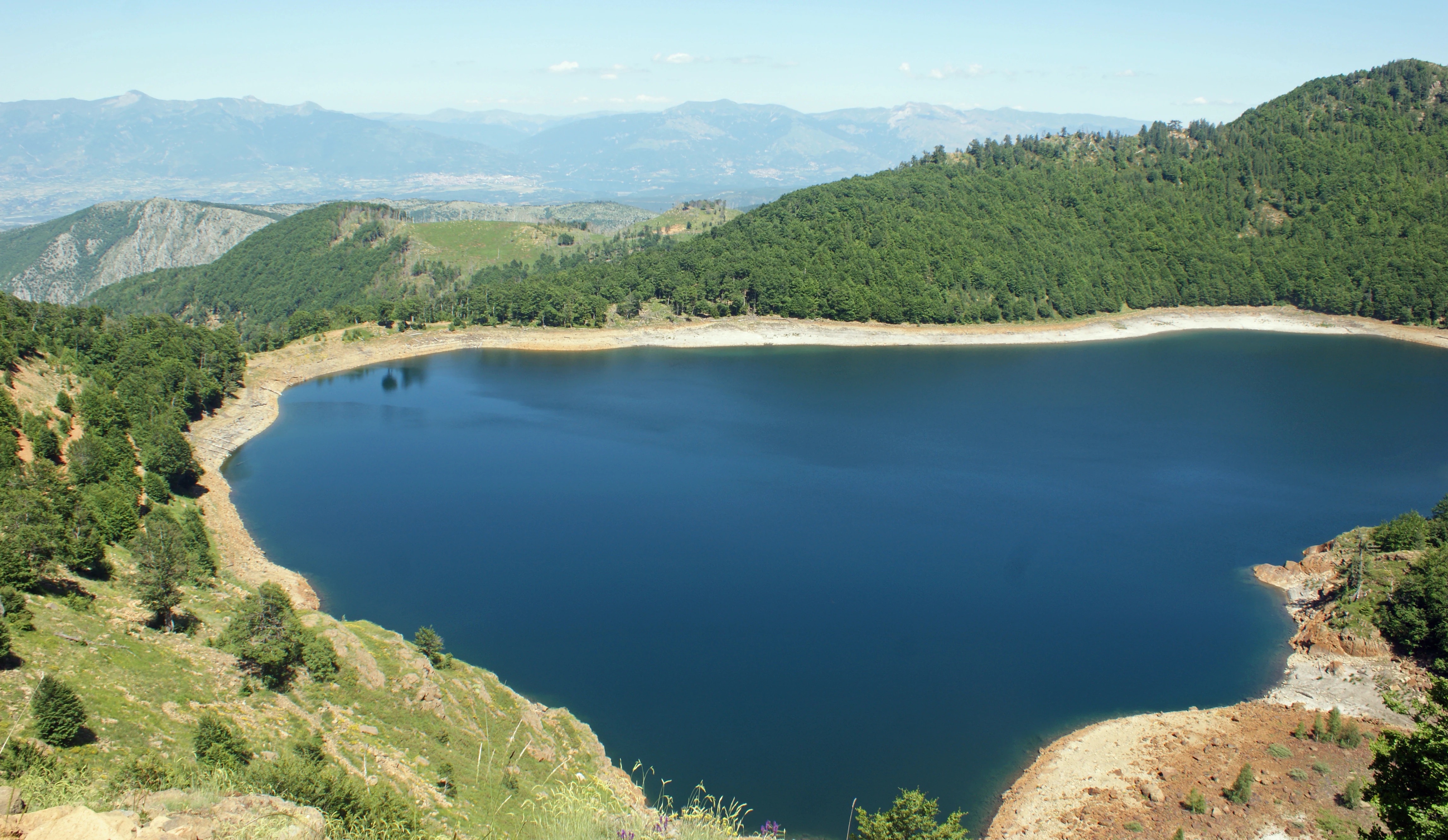 A high-angle view of a deep blue lake nestled within forested mountain slopes, showing clear water and rocky, grassy shorelines under a bright sky.