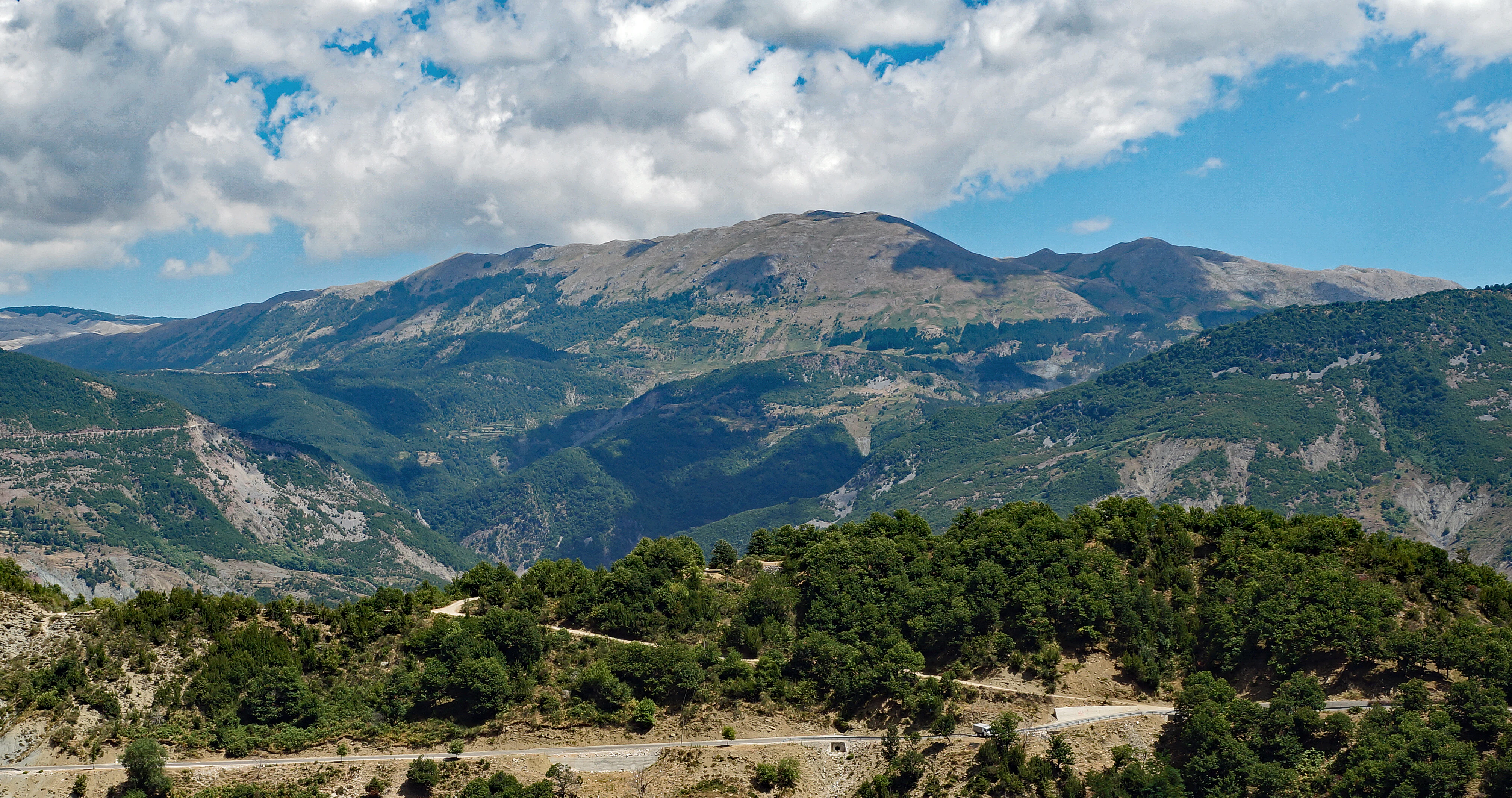 Panoramic view of rugged mountains and forested slopes in Mali me Gropa under a blue sky with scattered white clouds.