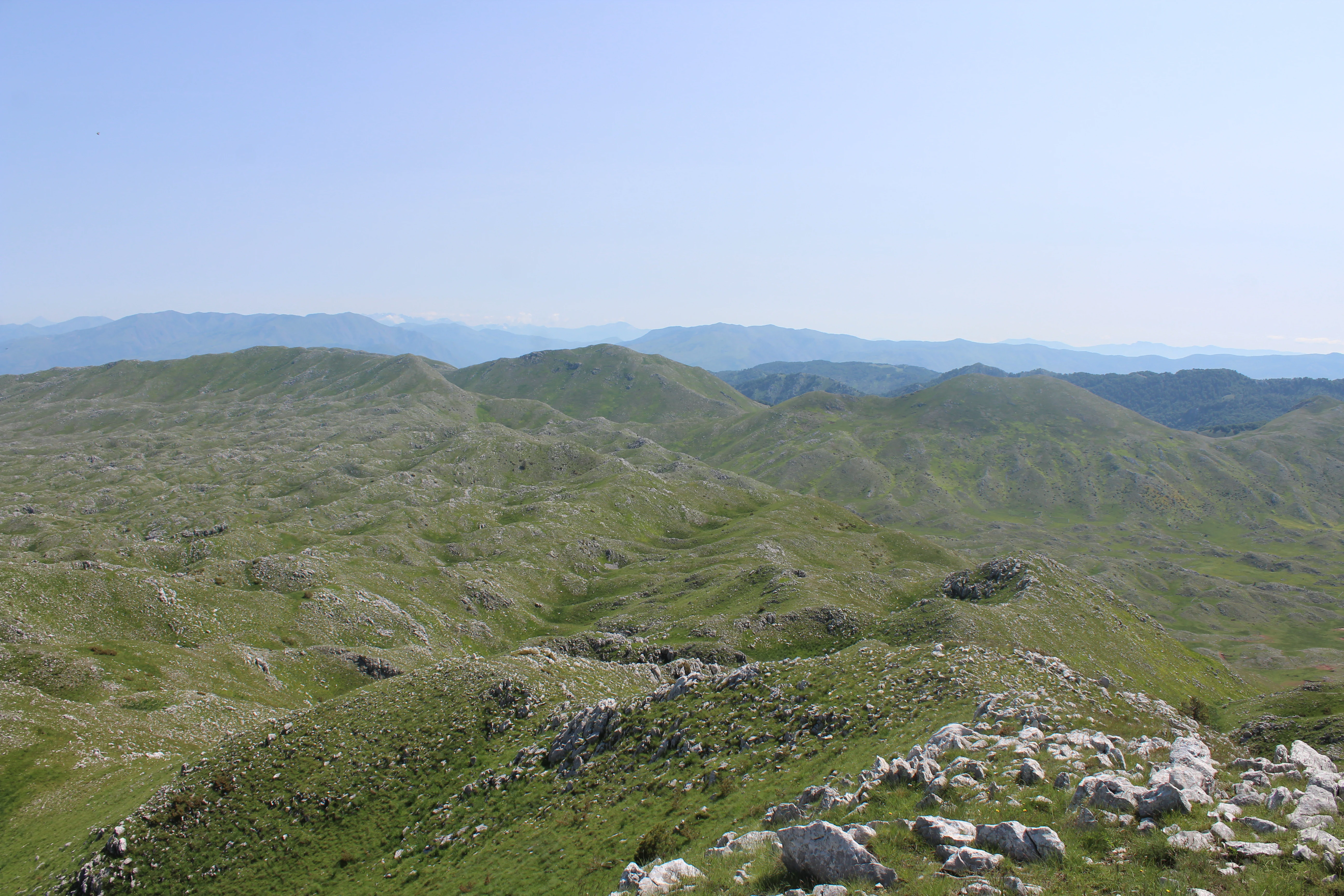 A wide, elevated view of undulating green hills and rocky, karst-like terrain under a clear blue sky, characteristic of the Albanian mountains.