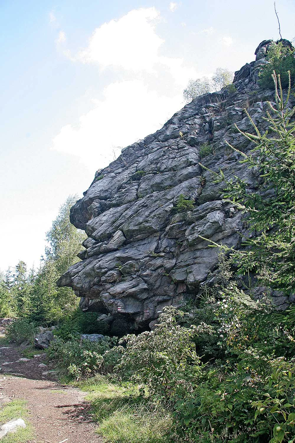 Una gran formación rocosa gris y escarpada se eleva sobre un sendero de tierra, con árboles de hoja perenne y vegetación escasa rodeando su base bajo un cielo parcialmente nublado.