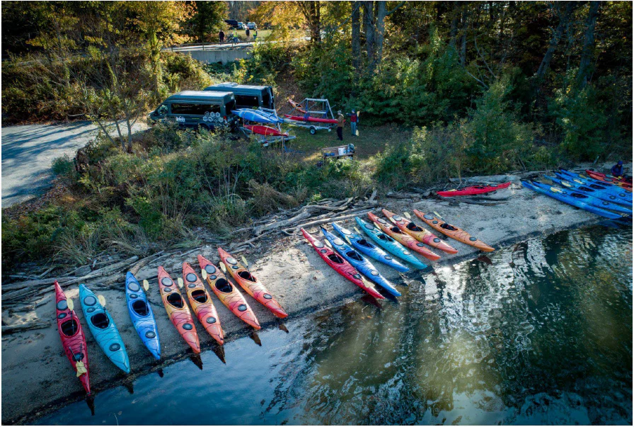 A high-angle view showing a row of many colorful kayaks lined up on a narrow, sandy beach next to a river with a wooded bank in the background.