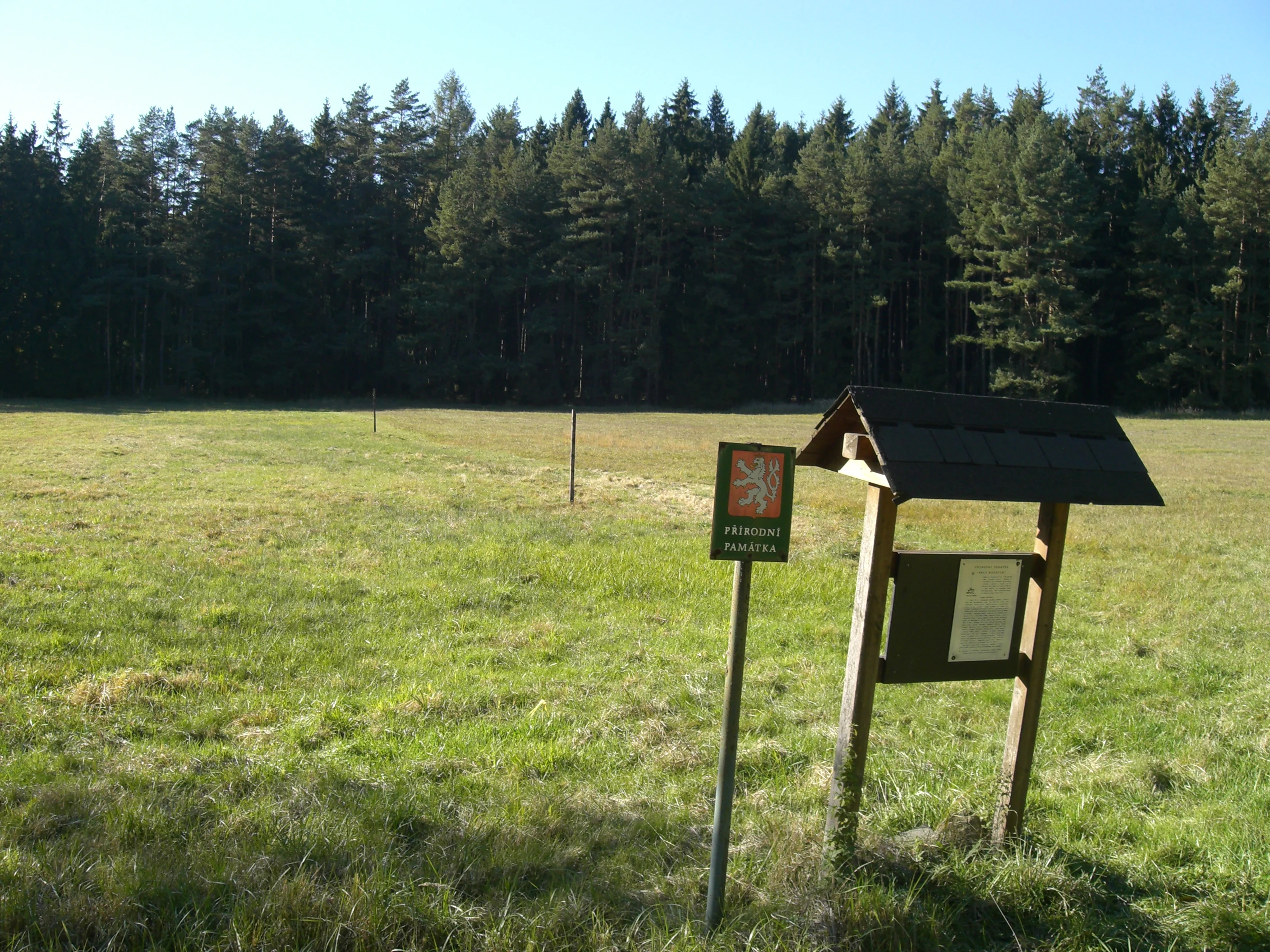 A wooden information sign and a small shield displaying the Czech national emblem stand in a grassy meadow bordered by a pine forest.