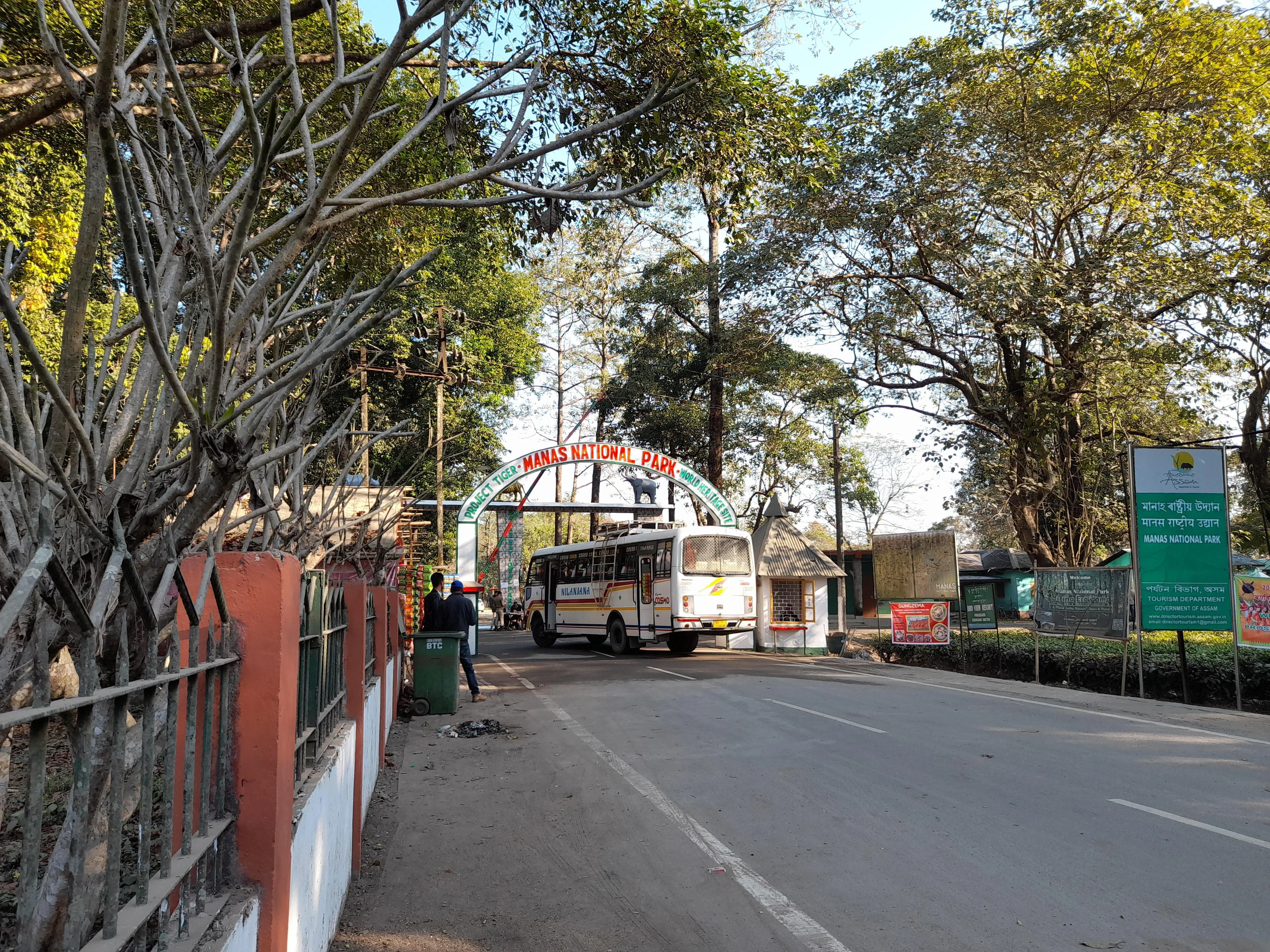 Entrance gate structure with 'Manas National Park' signage, a bus on a paved road, trees, and green informational signs