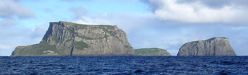 A wide shot from the ocean showing two steep, flat-topped rocky islands with sheer cliffs rising above the deep blue water under a cloudy sky.