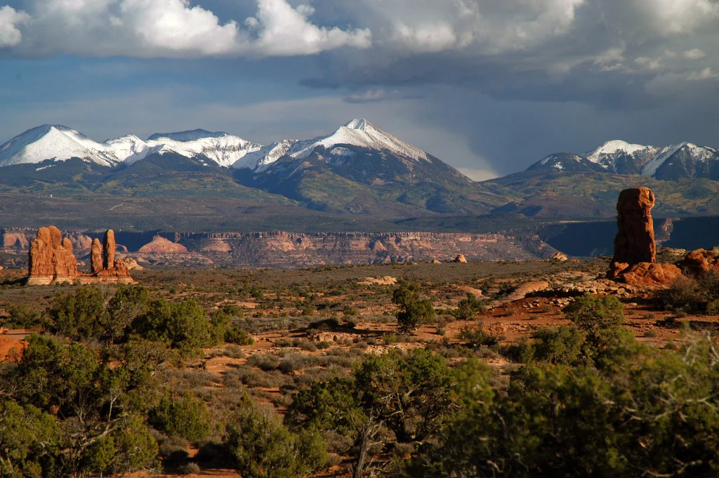 A distant view of snow-covered mountain peaks rising above a desert landscape with red rock formations and scrub brush in the foreground.