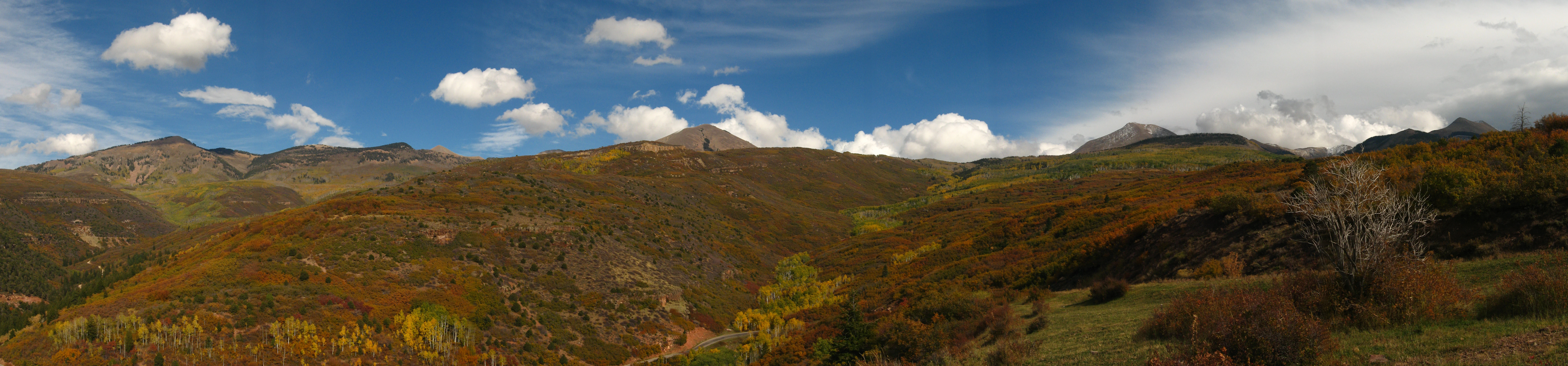 A wide panoramic view of the La Sal Mountains featuring rolling slopes covered in fall foliage under a blue sky with scattered clouds.