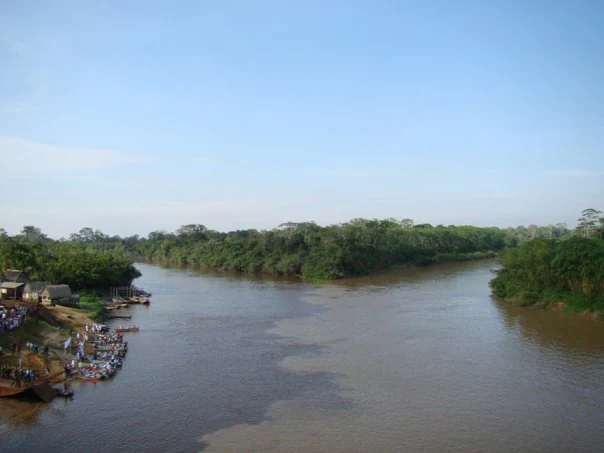 An aerial-style view of a river confluence in a lush, green Amazonian landscape, with a small gathering of boats docked at a riverbank on the left side.