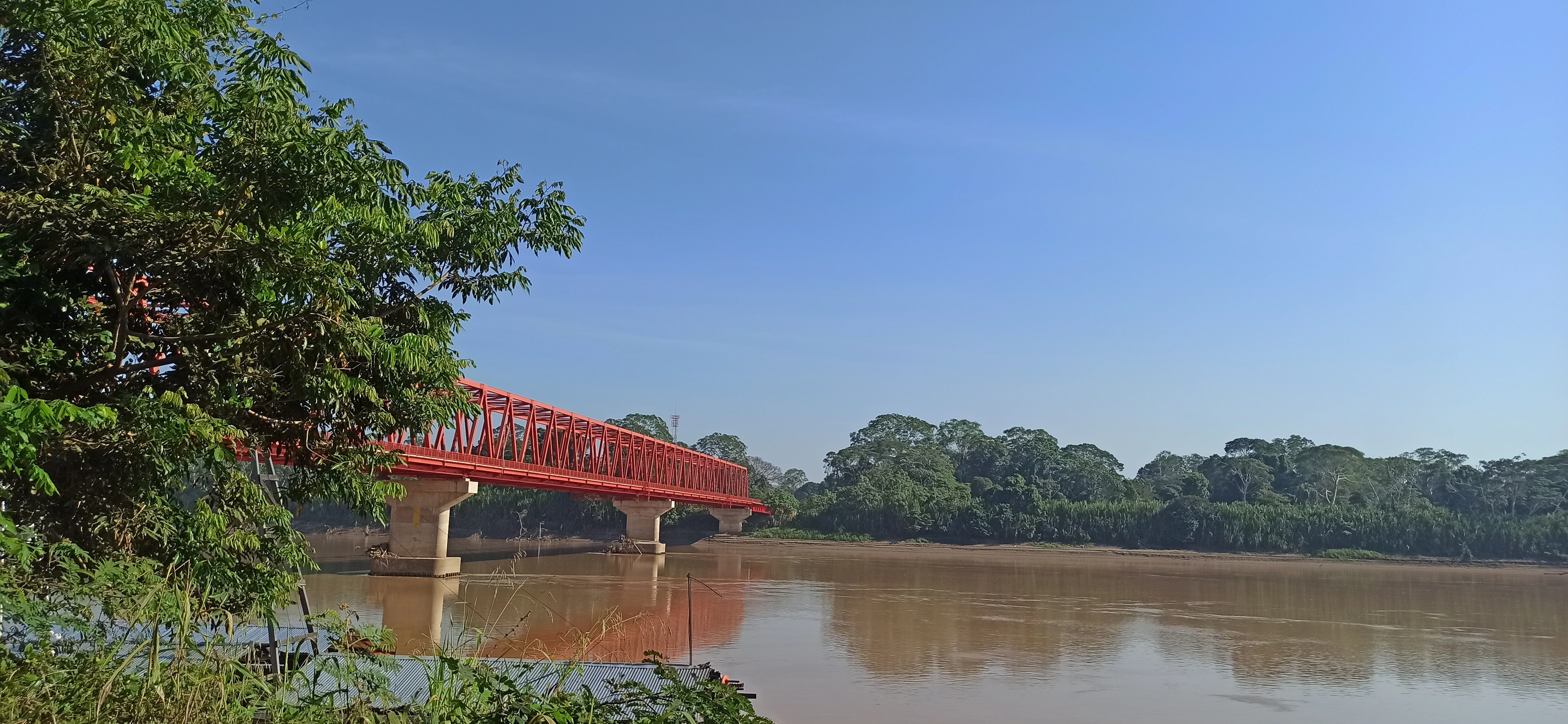 A long, bright red metal truss bridge crosses a wide, silt-heavy brown river under a clear blue sky, surrounded by lush green Amazonian forest on both banks.