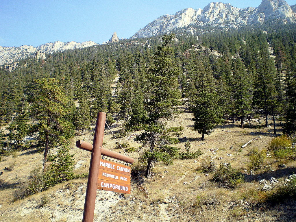 A brown wooden park sign reading Marble Canyon Provincial Park Campground, situated in a dry, coniferous forest with high limestone cliffs in the background.