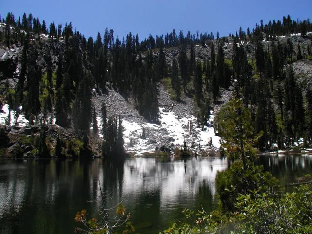 A calm lake reflecting snow-patched rocky ridges and a dense coniferous forest under a clear blue sky.