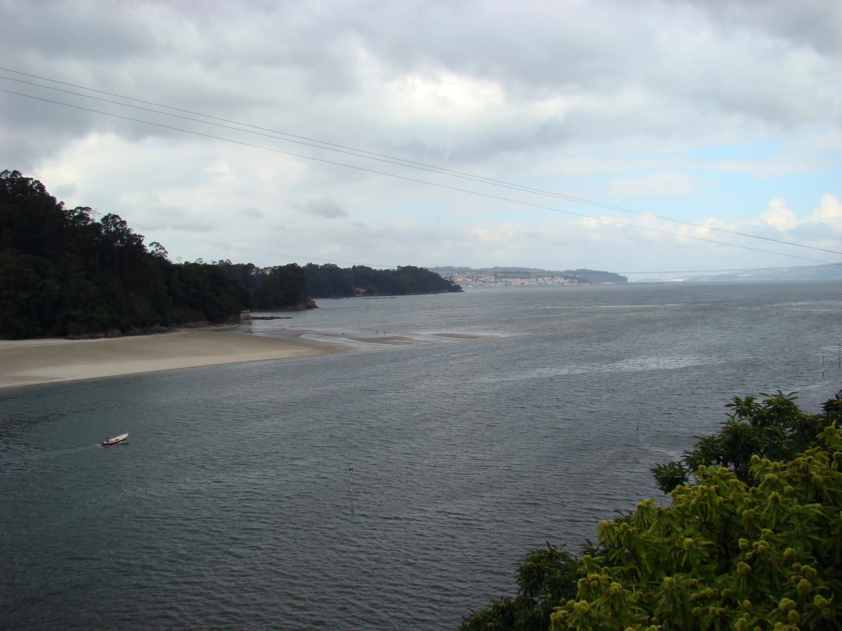 A wide view of a river estuary under a cloudy sky, showing a sandy beach on the left, calm dark water, and dense green shoreline vegetation.