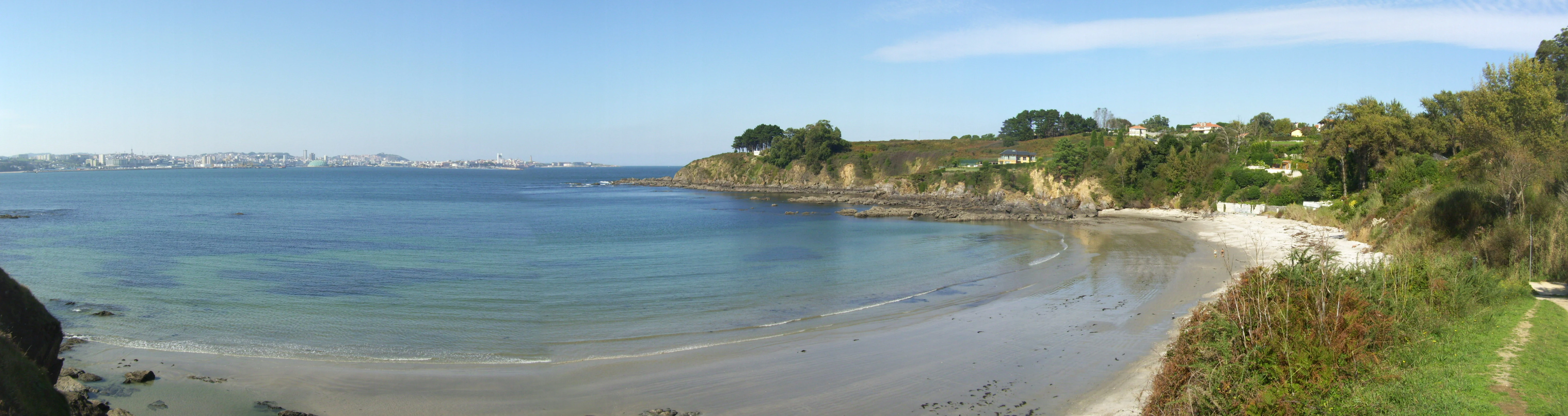 A wide panoramic view of a sandy beach in a cove with calm water, grassy cliffs on the right, and a hazy city visible on the far horizon.