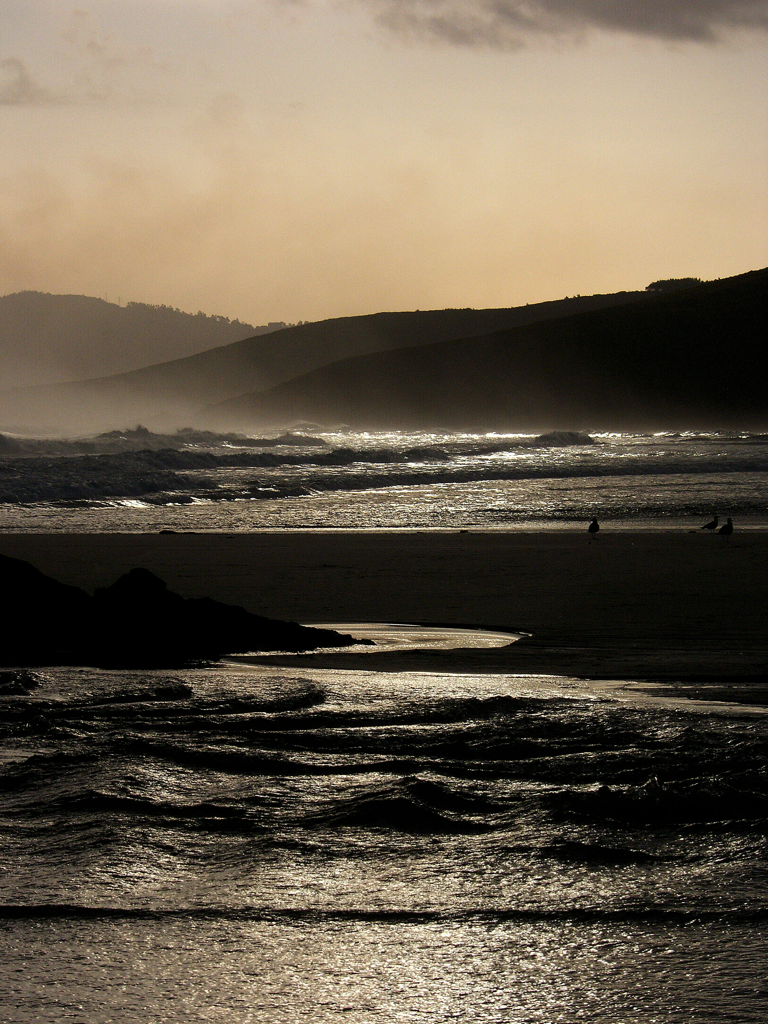 A backlit coastal landscape in Galicia at sunset, showing dark rolling hills in the background, a sandy beach, and shimmering dark water in the foreground.