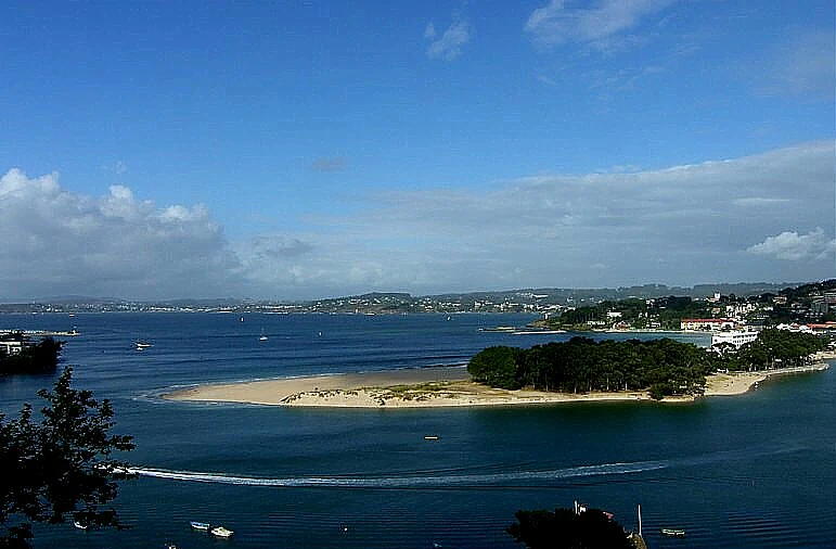 A wide view of Santa Cristina Beach, featuring a sandy spit of land surrounded by calm blue water, with coastal vegetation and buildings visible in the distance under a sunny, partly cloudy sky.