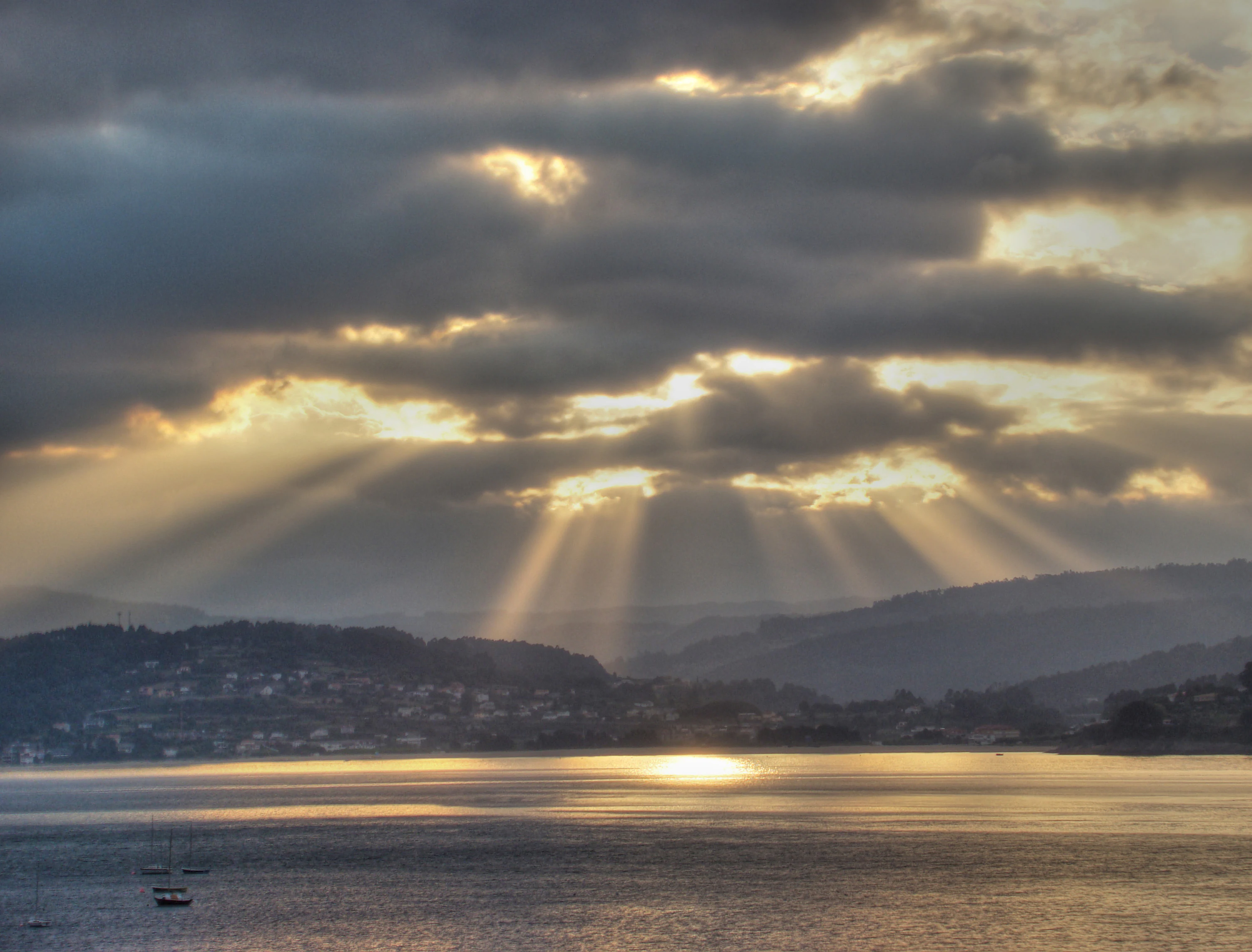 Sunbeams break through heavy clouds at sunrise over a calm coastal bay with distant hills and a small town.