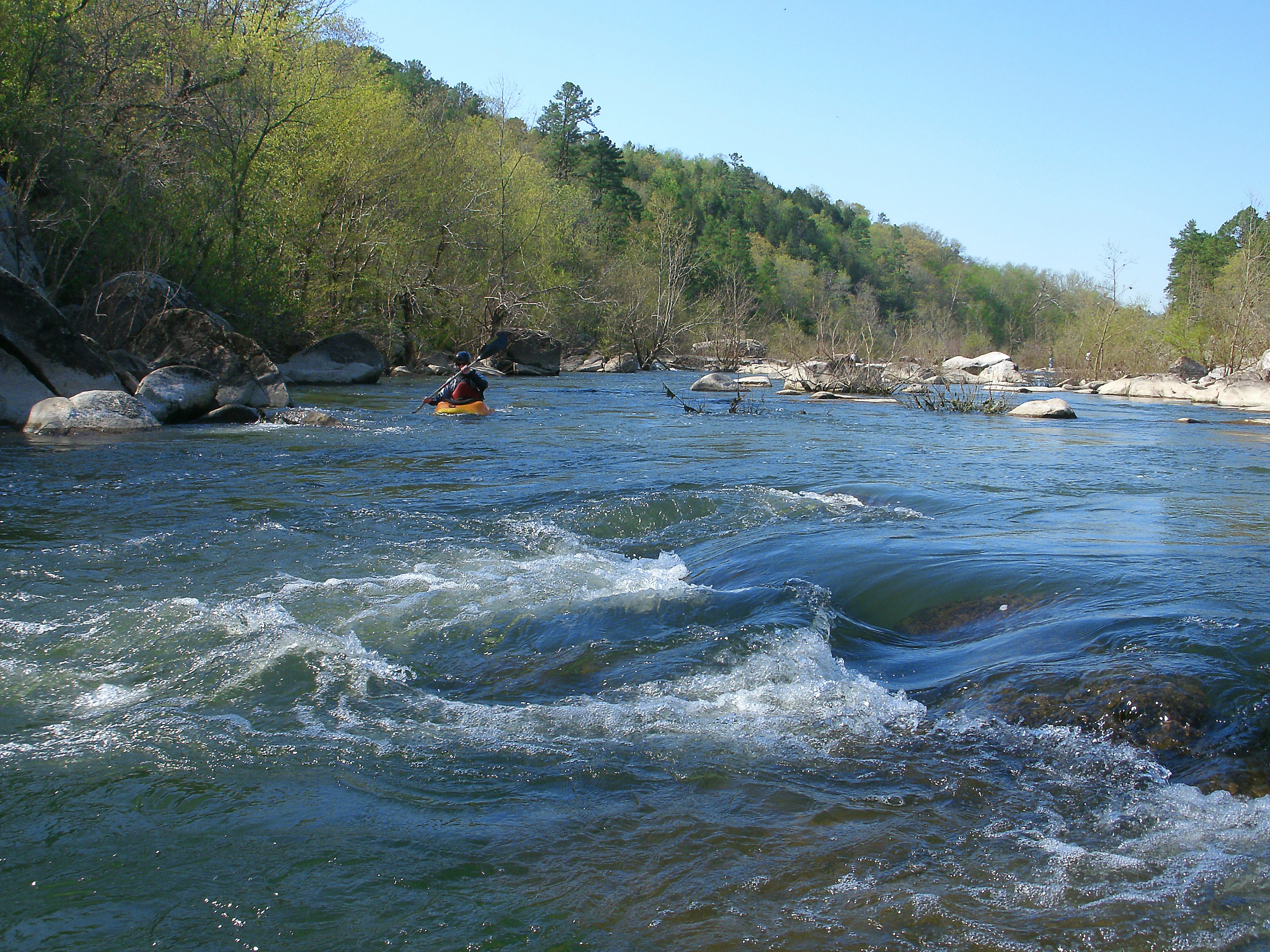 A person in a yellow kayak paddling down a river with rocky banks and a forested hillside in the background.