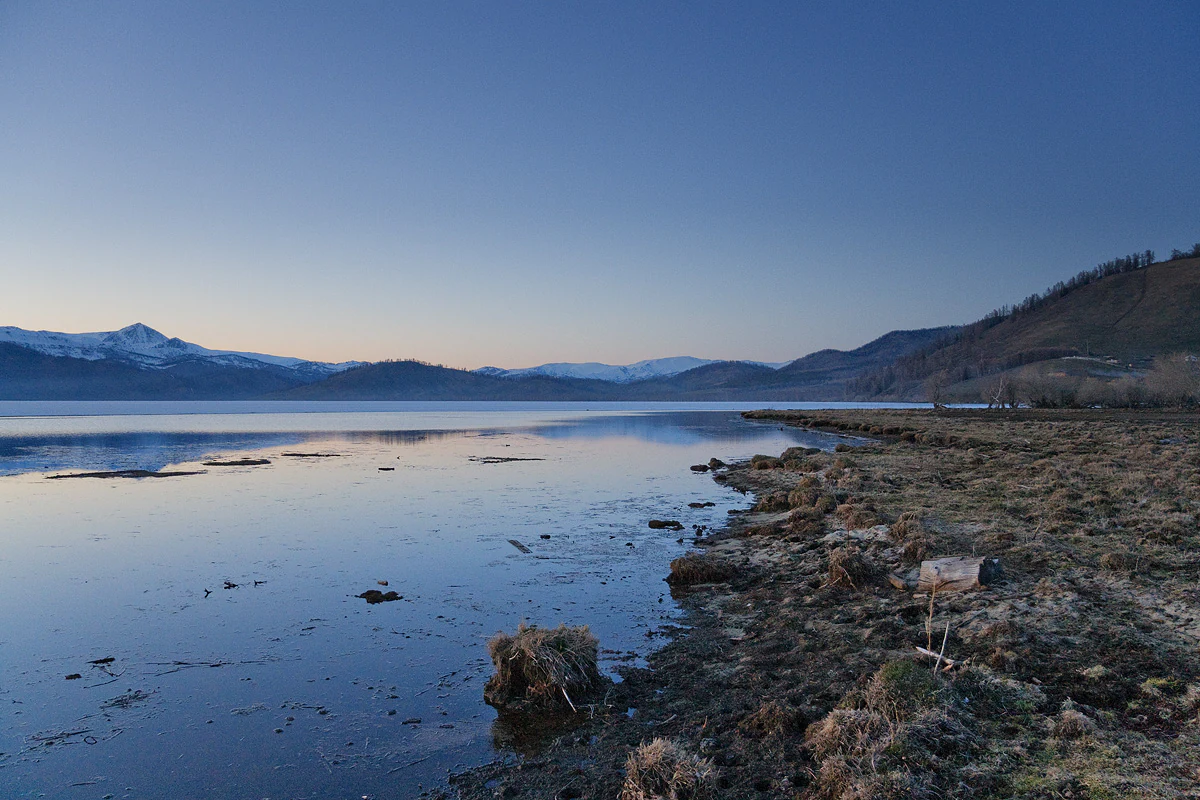 A calm, reflective lake with a shoreline of low, sparse vegetation in the foreground and snow-dusted mountain ridges visible under a clear, cool-toned sky.