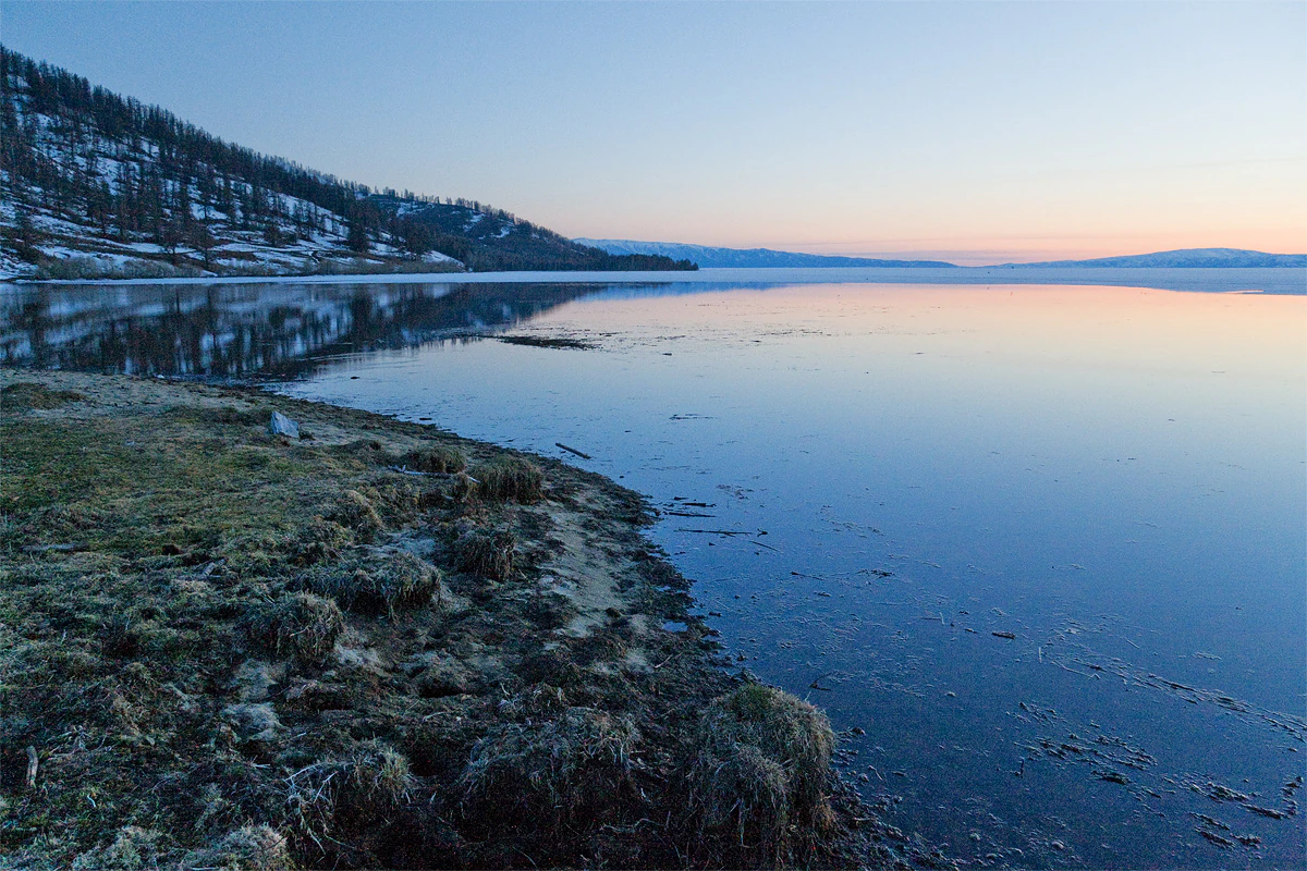 A peaceful, wide landscape view of Lake Markakol at dusk, with snow-dusted hills in the background reflected in the calm, dark blue water of the lake. The foreground features a rugged, grassy shoreline.