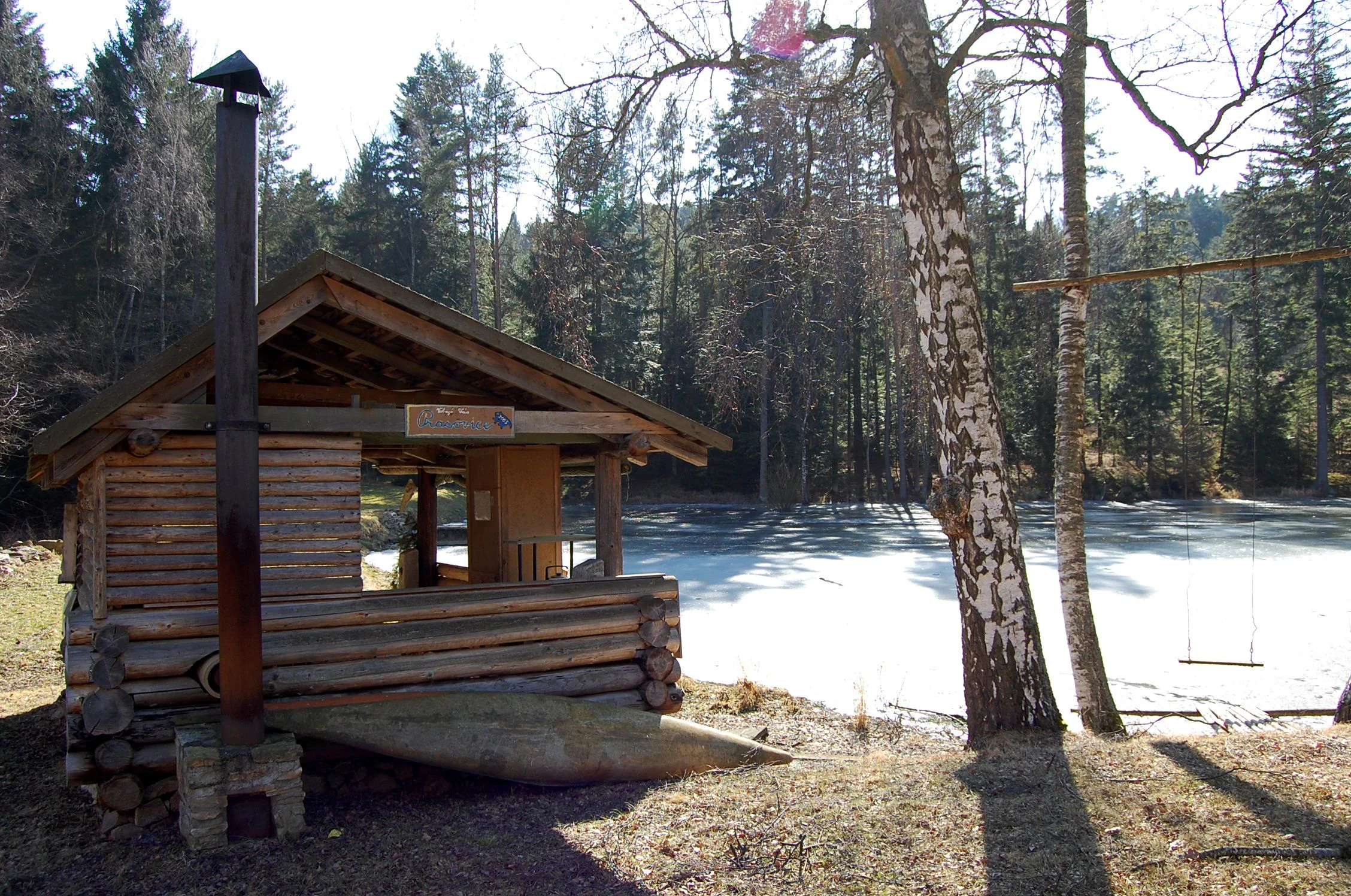A small rustic wooden cabin with a chimney sitting next to a partially frozen pond, surrounded by a forest with a prominent birch tree in the foreground.