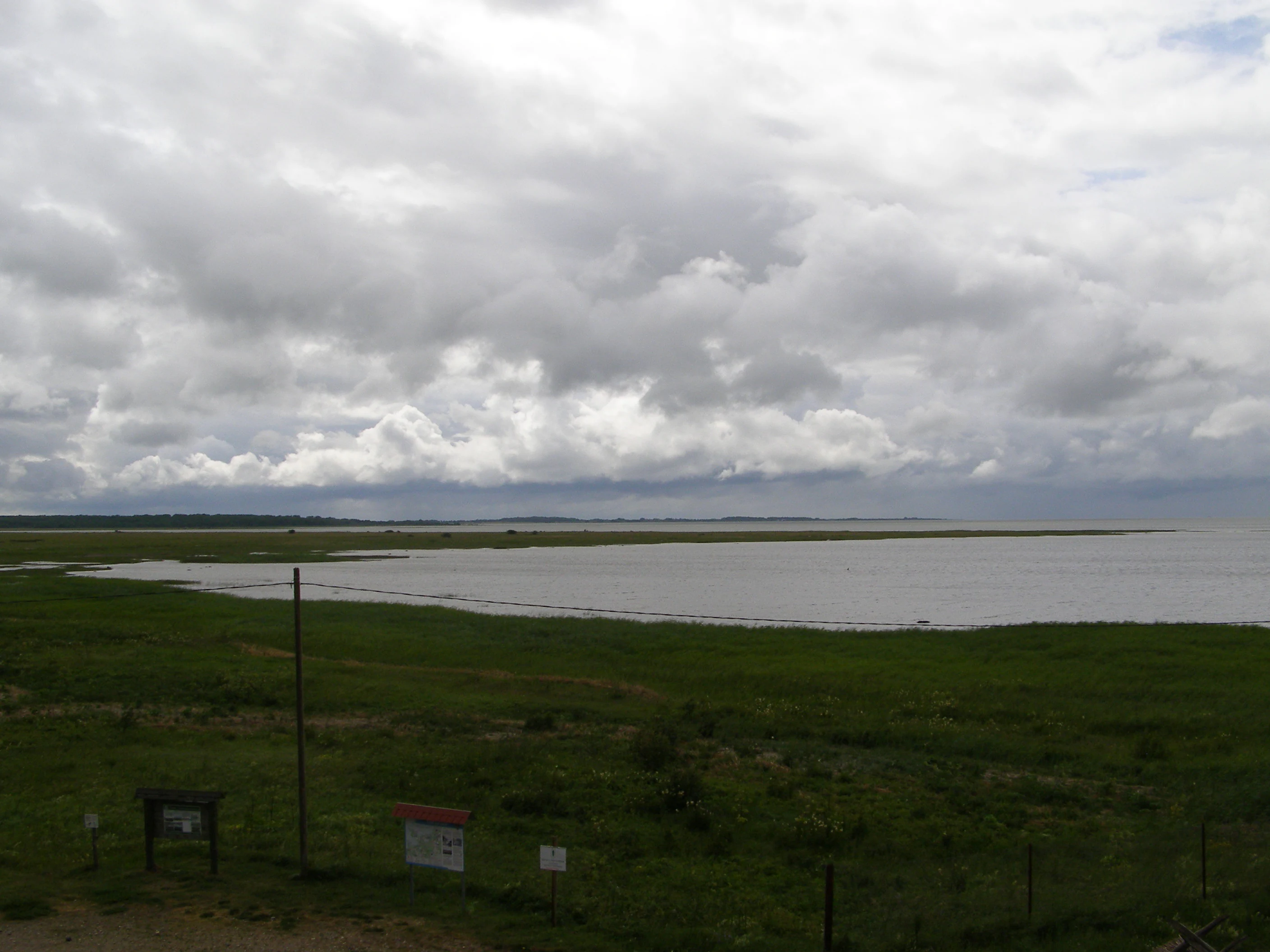 Grassy field with informational signs overlooking a large body of water under a cloudy sky