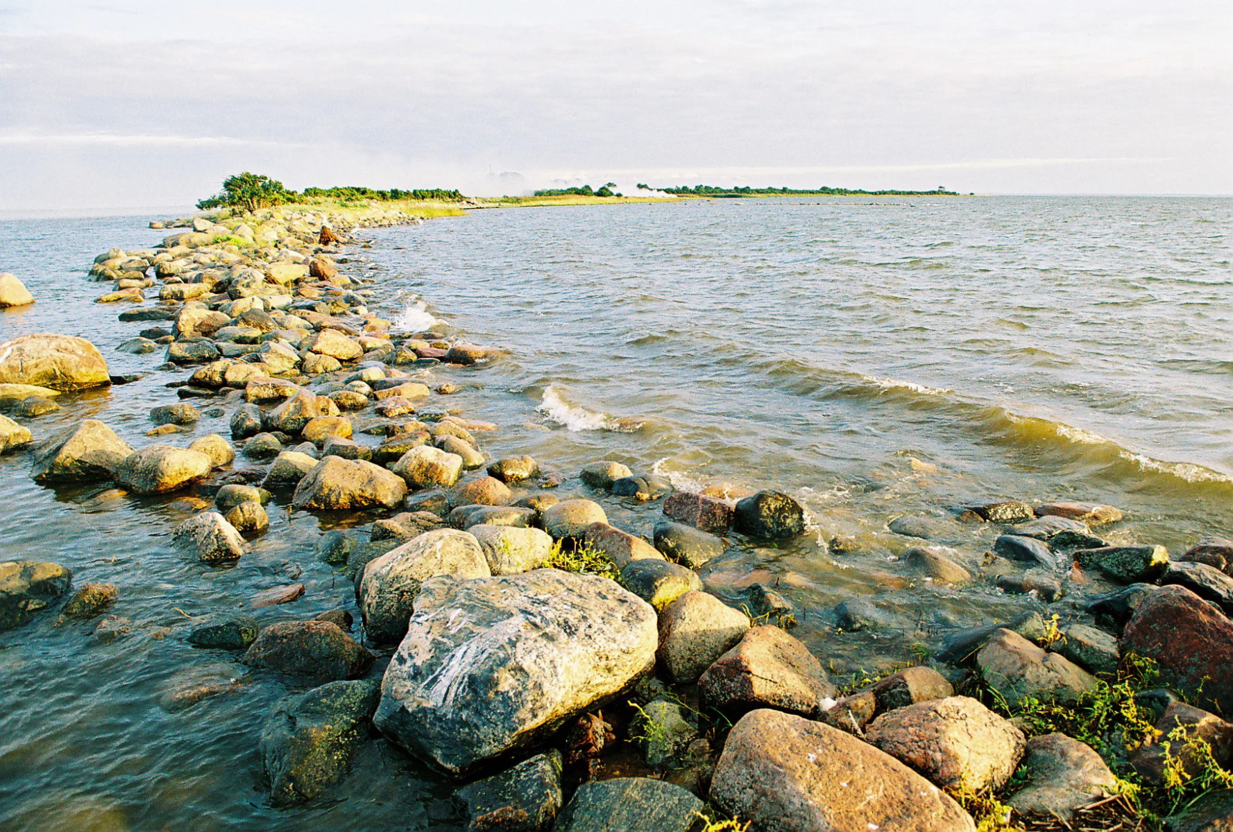Rocky shoreline with stone jetty extending into calm water, distant islet with vegetation under partly cloudy sky
