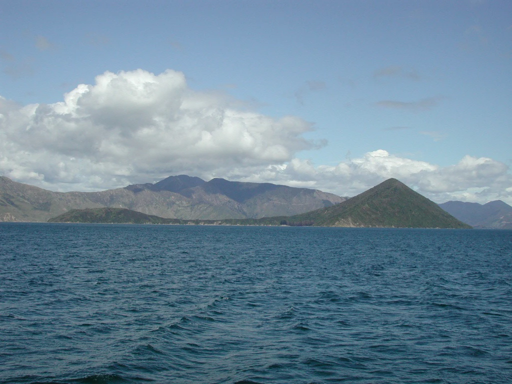 Una vista gran angular de una isla verde y montañosa rodeada de aguas azul profundo bajo un cielo parcialmente nublado.