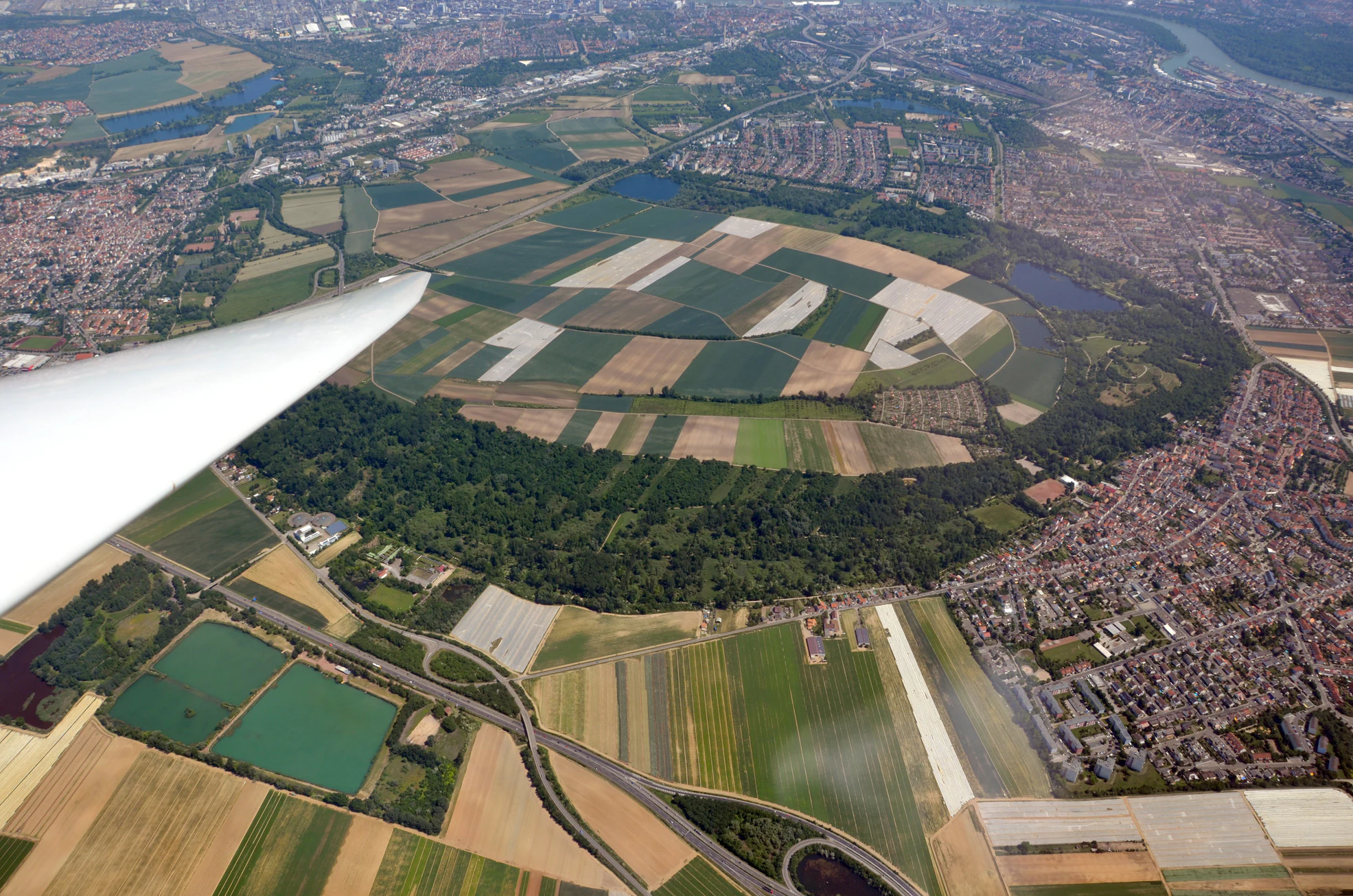 Vista aérea desde un planeador que muestra campos agrícolas y una zona central de humedal boscoso rodeada de viviendas suburbanas.