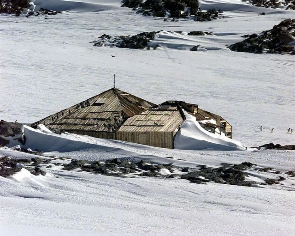 A wooden, pyramid-roofed structure sitting in a vast, snow-covered landscape under a bright sky, characteristic of the historic Mawson's Huts site in Antarctica.