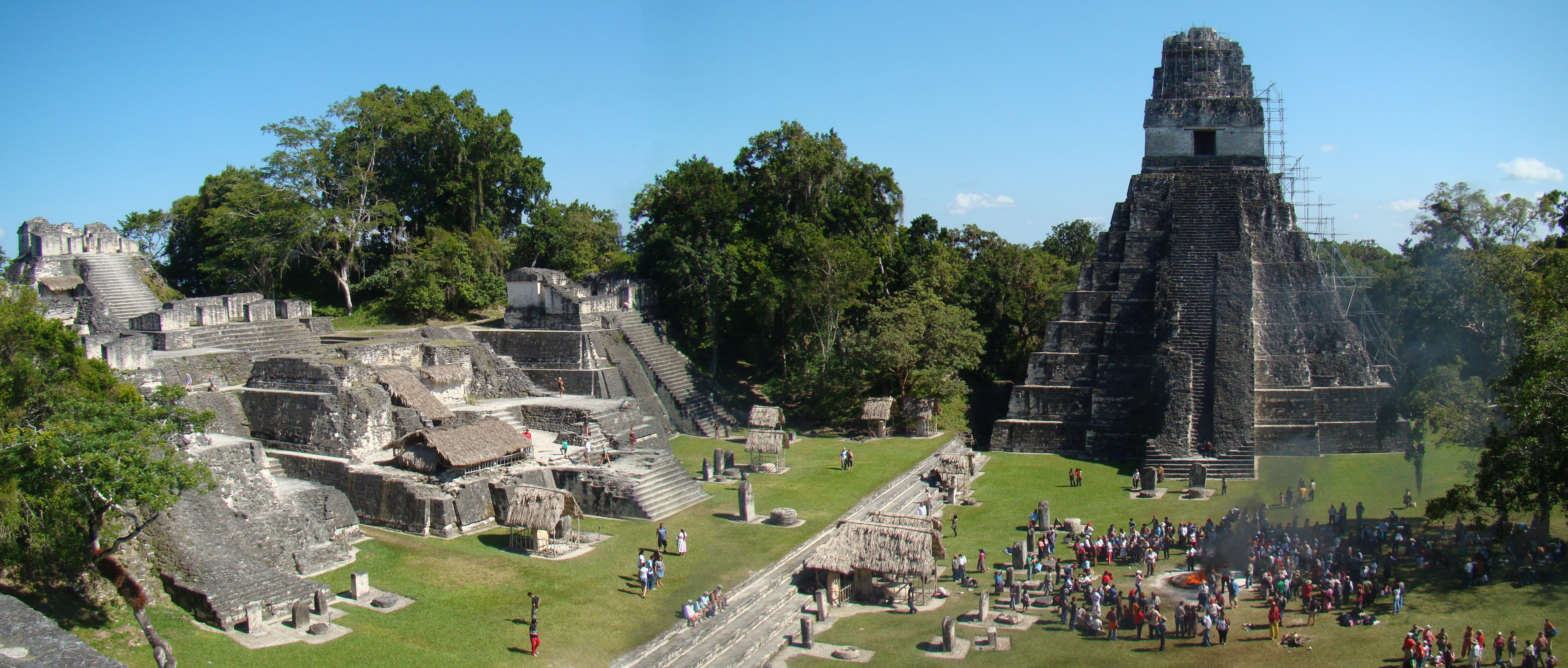 A panoramic wide shot of the Tikal Great Plaza, featuring a tall stone temple on the right, the North Acropolis ruins on the left, and a large crowd gathered in the central grassy plaza.