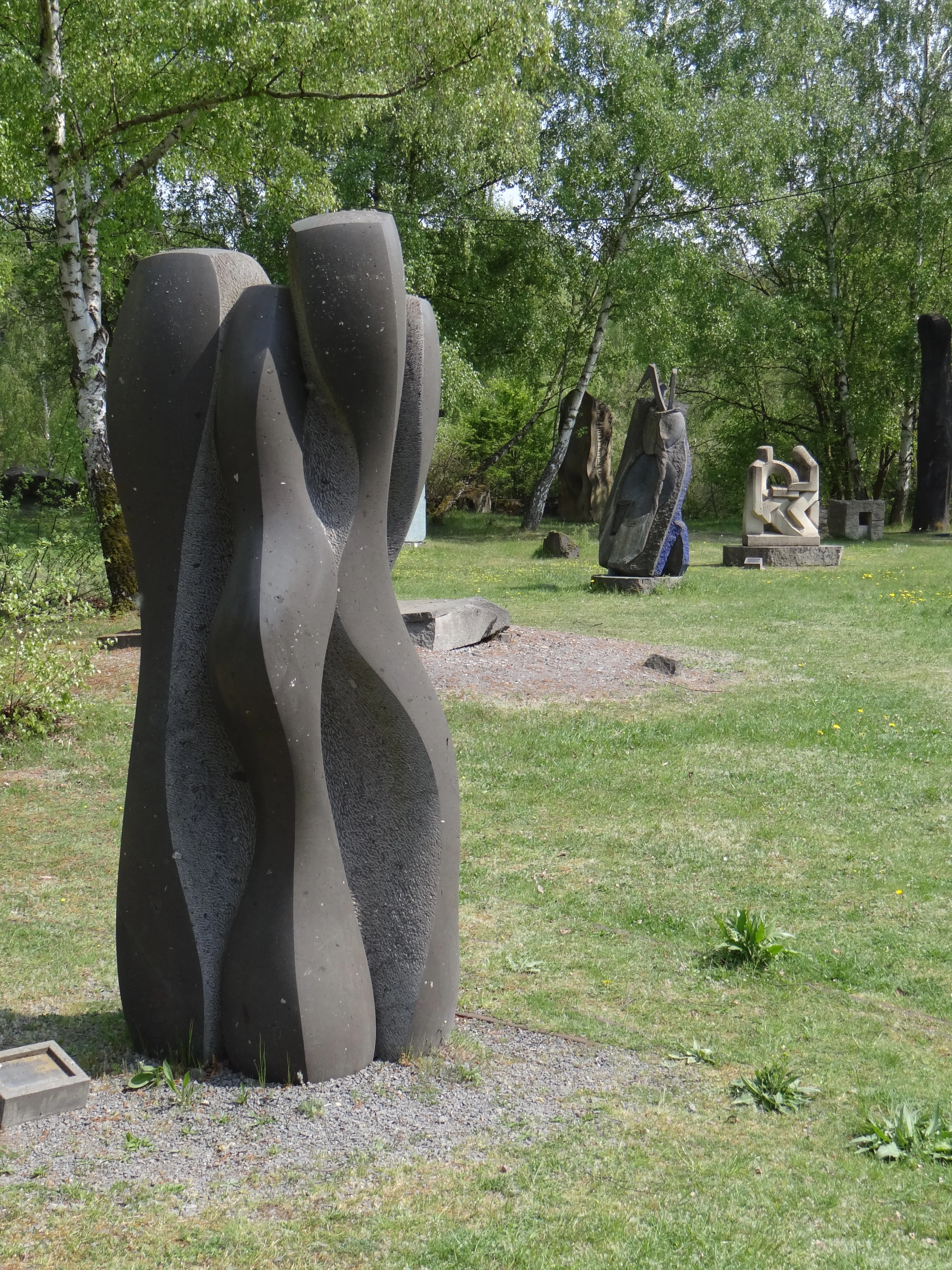 A large, dark, abstract stone sculpture stands in the foreground, with several other sculptures placed on a grassy lawn surrounded by trees.