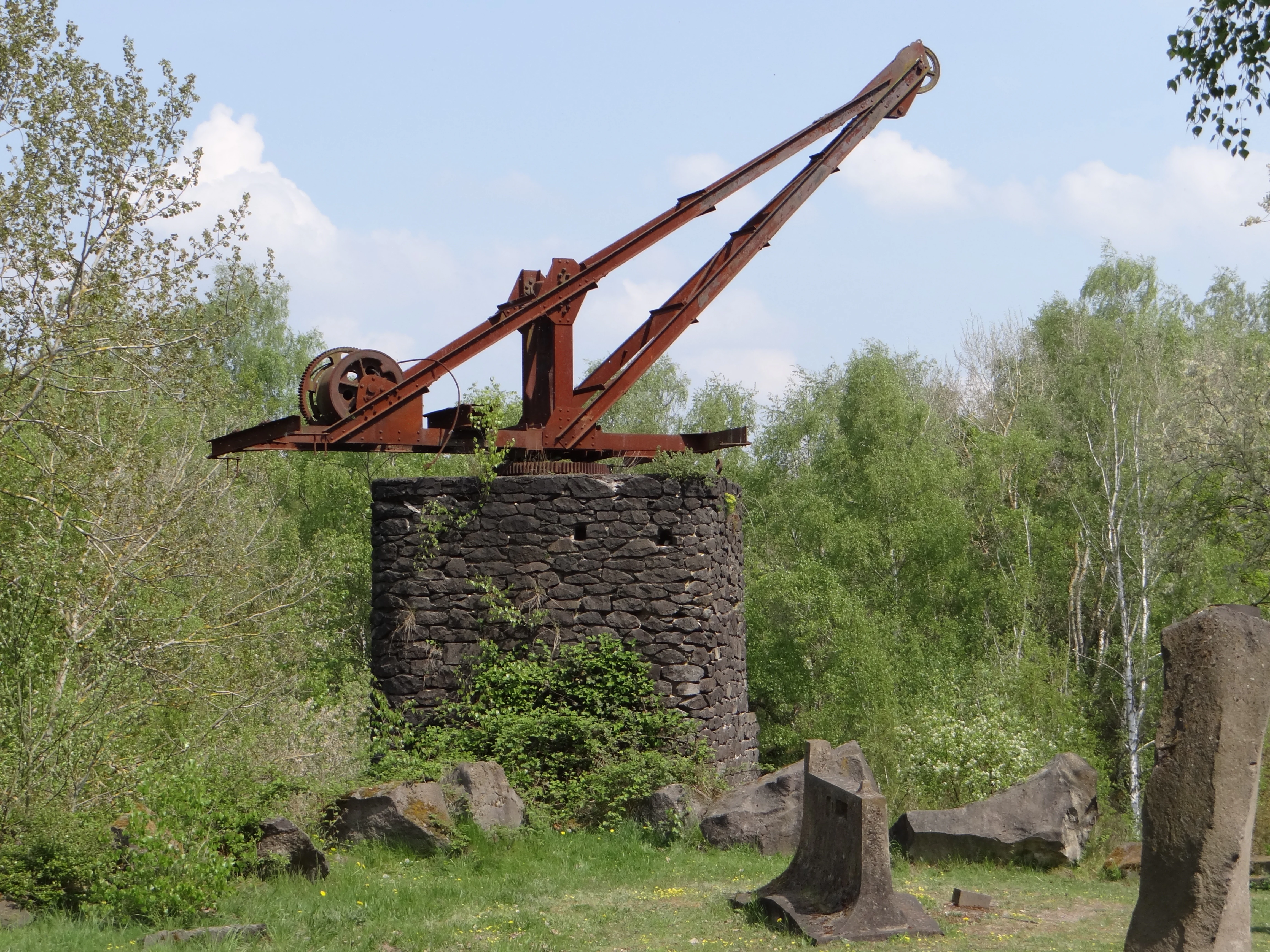 A weathered, rusted historic quarry crane sits atop a circular dry-stacked stone pedestal, surrounded by green trees and scattered basalt boulders.