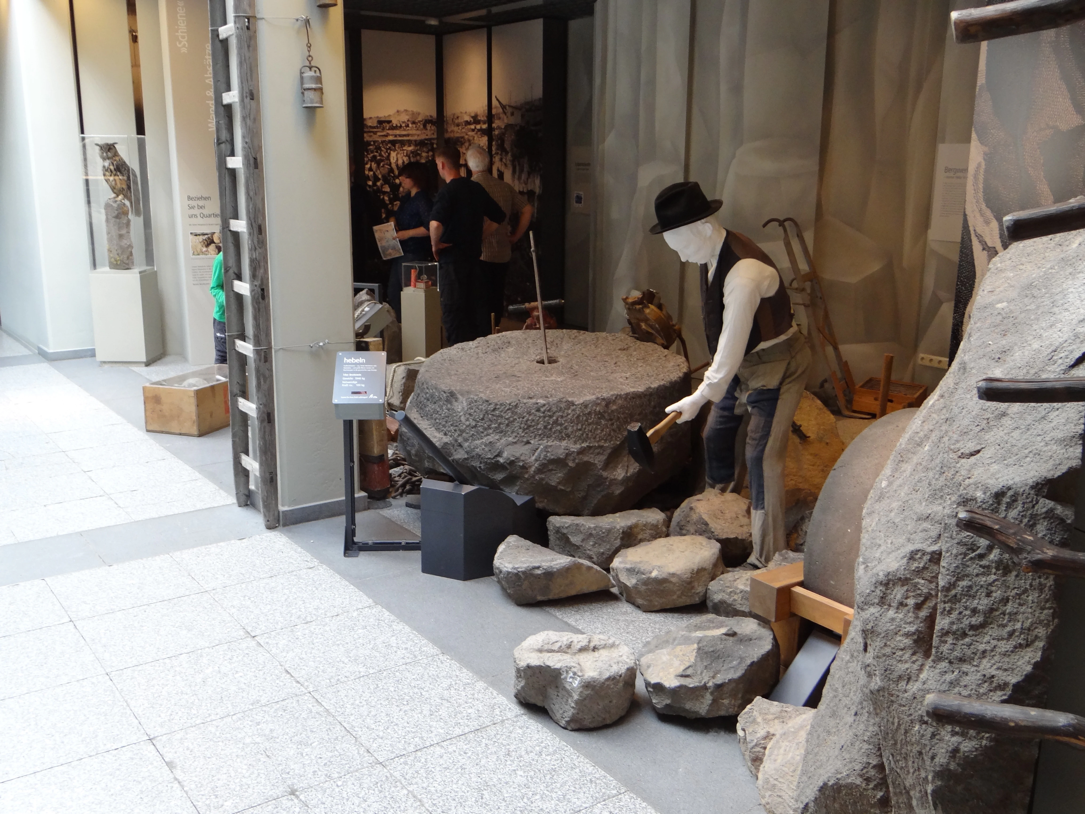 A life-sized diorama in a museum featuring a mannequin dressed in historic work clothing, holding a hammer over a large circular basalt stone, representing traditional quarrying activities.