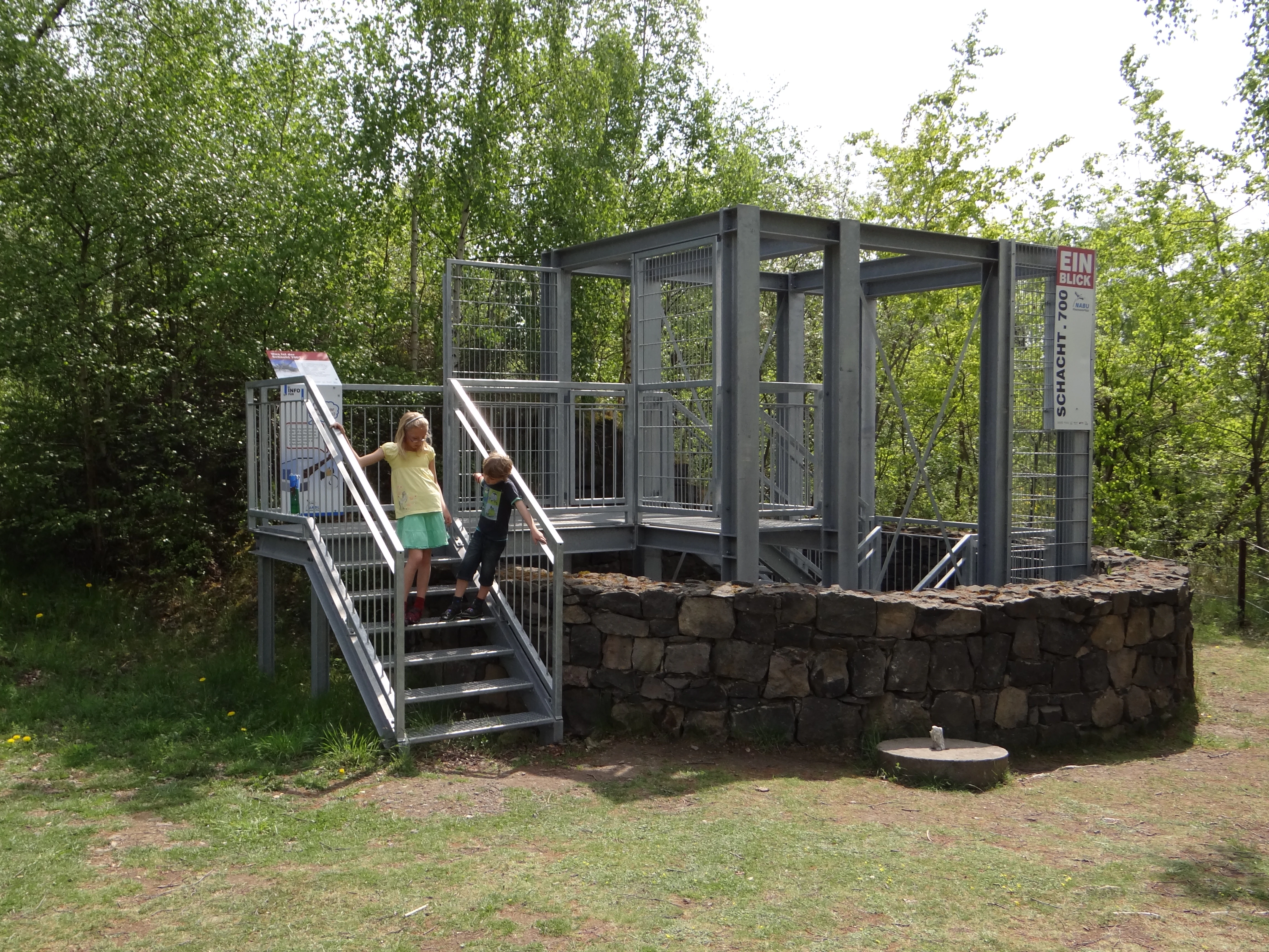 A metal viewing platform and fenced entrance structure over a mining shaft, with two children standing on the metal stairs, surrounded by trees and a stone retaining wall.