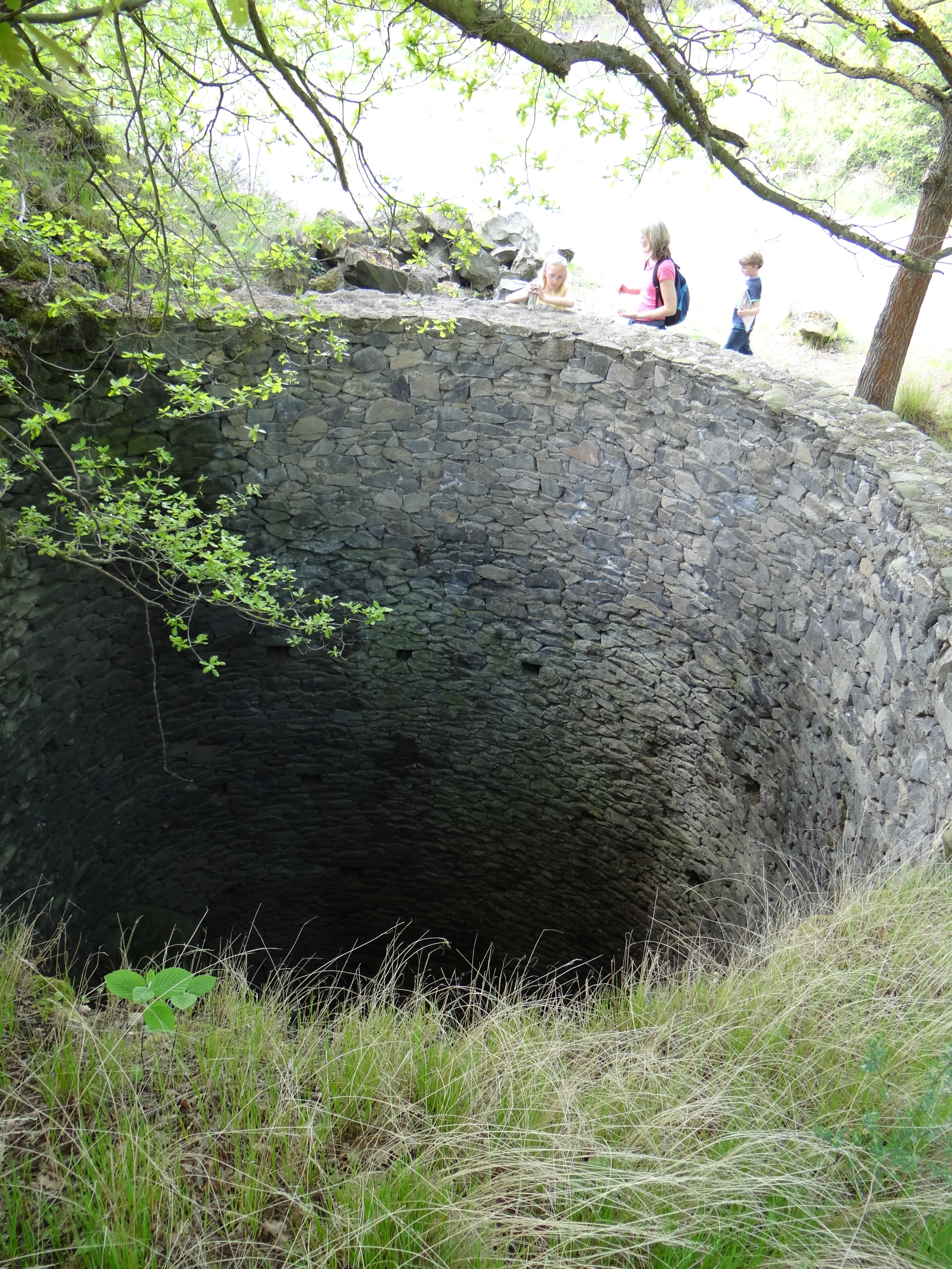 A deep circular stone shaft at the Mayener Grubenfeld park, with a stone retaining wall and visitors walking in the background.