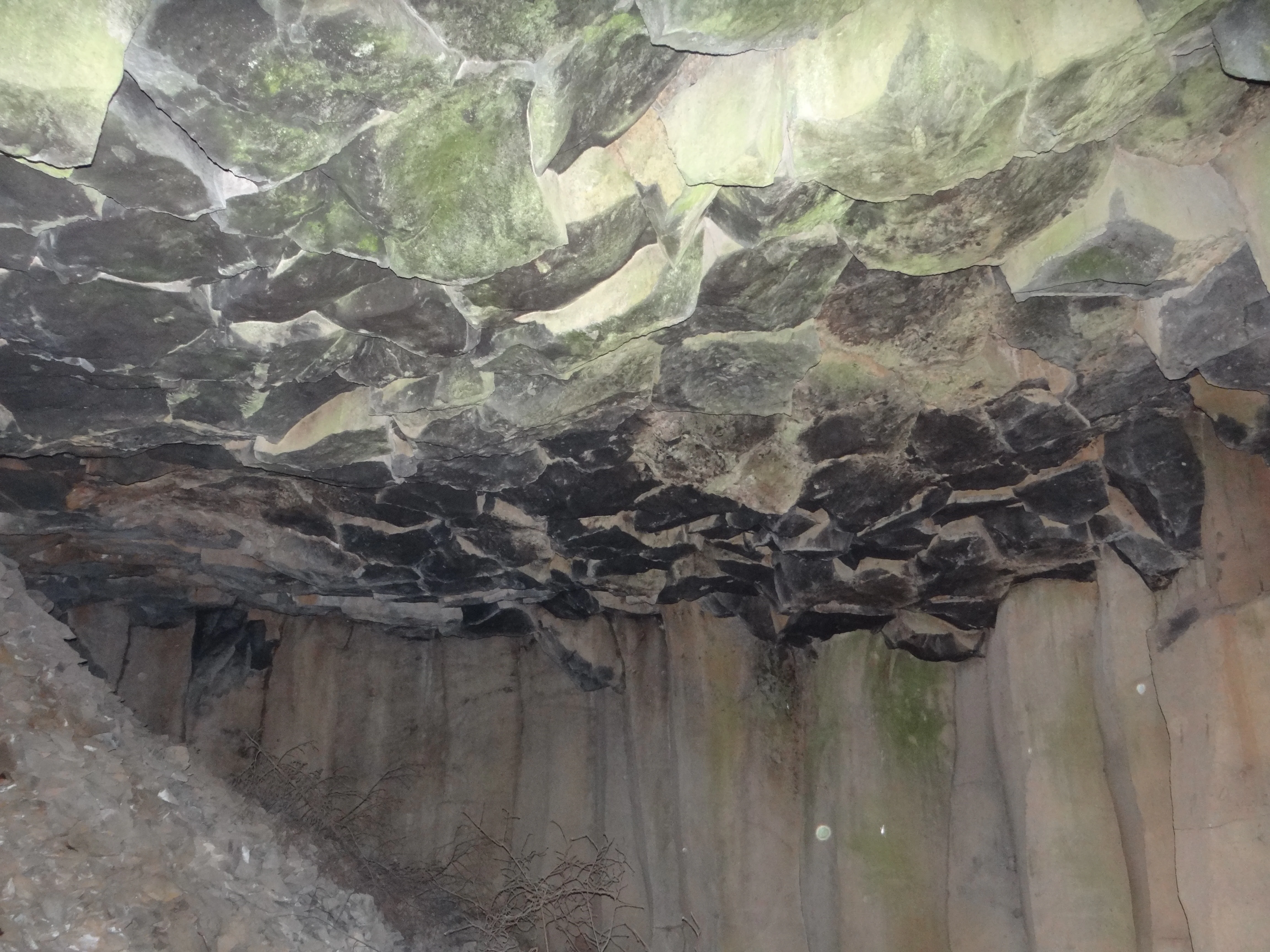 A close up view of dark, hexagonal basalt column formations creating a natural ceiling above a rocky quarry wall.