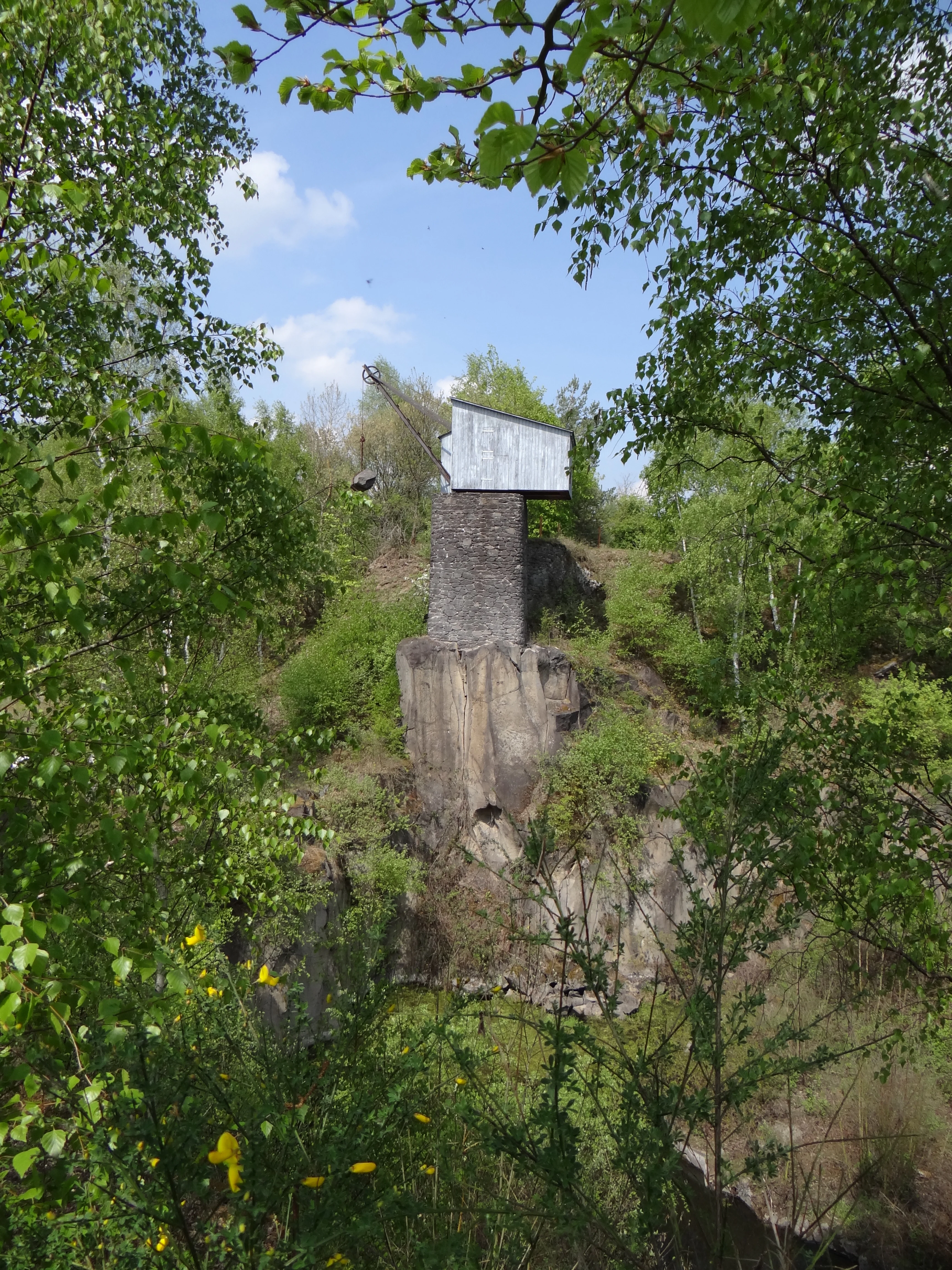 A vintage crane with a stone base and metal housing perched on a rock formation within a quarry, framed by birch trees.