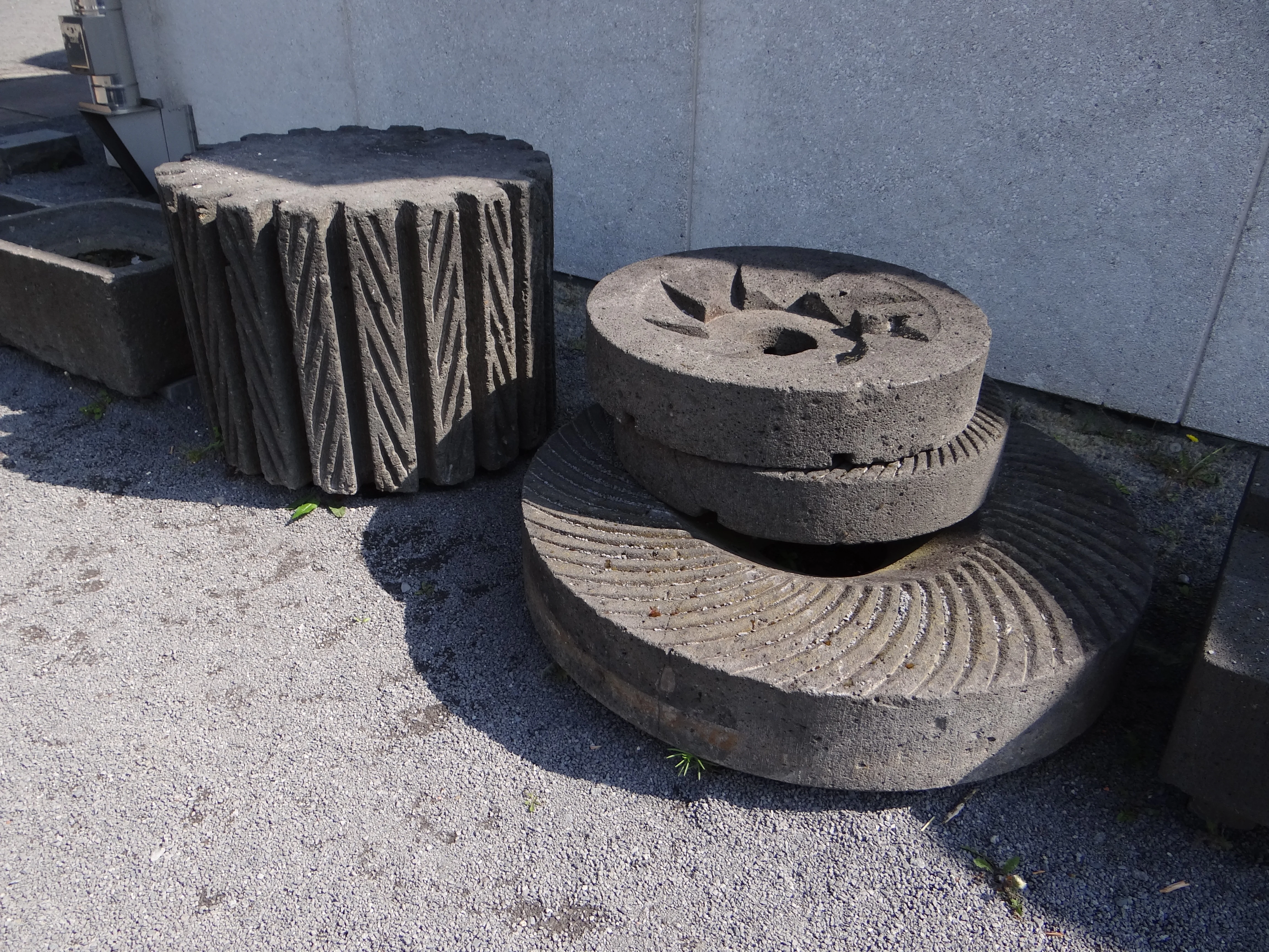 A collection of carved circular basalt millstones resting on a gravel surface next to a white wall.