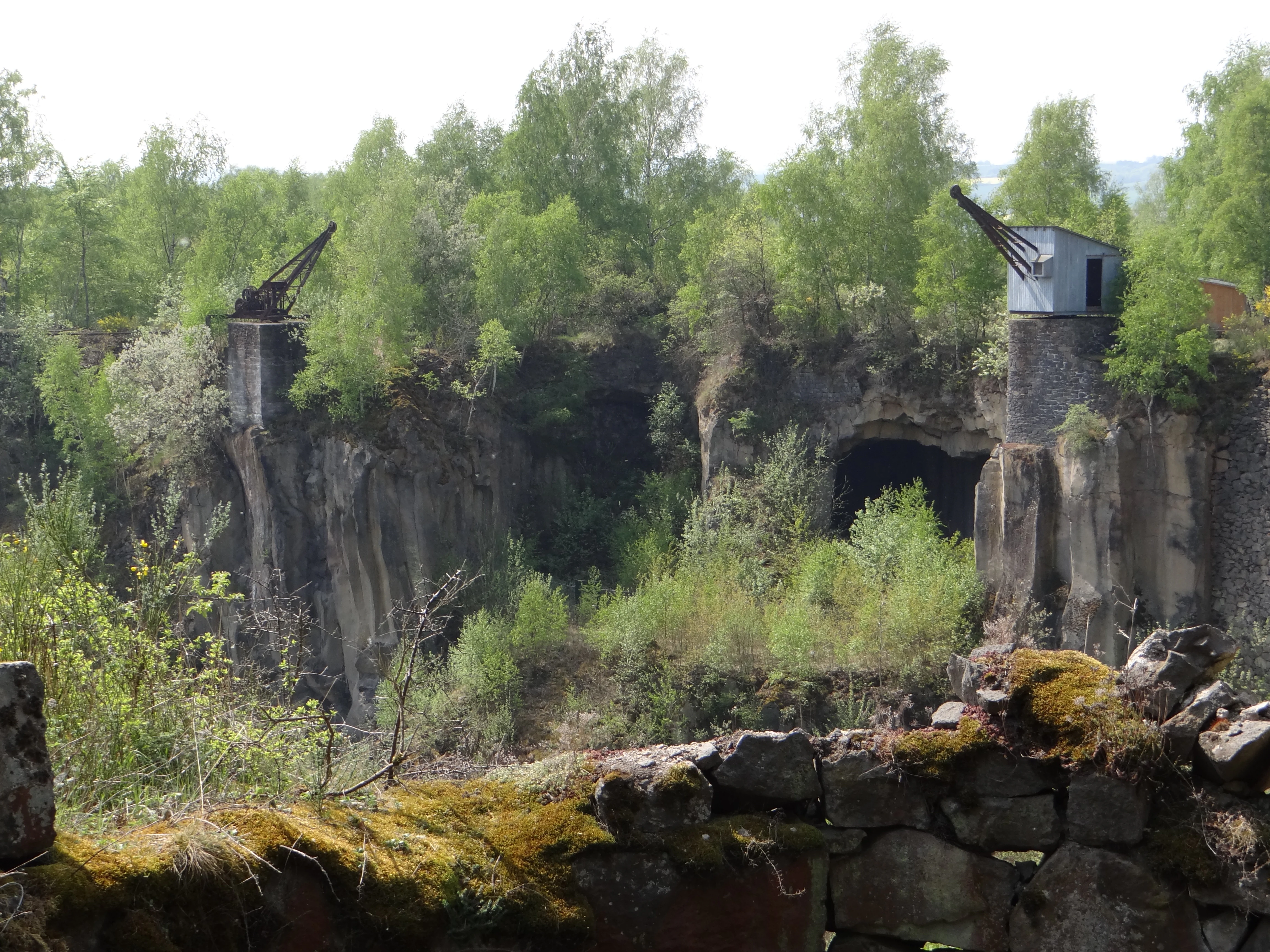 A view of the Mayener Grubenfeld quarry showing rocky cliff faces, deciduous trees, and two old industrial cranes perched on stone pillars.
