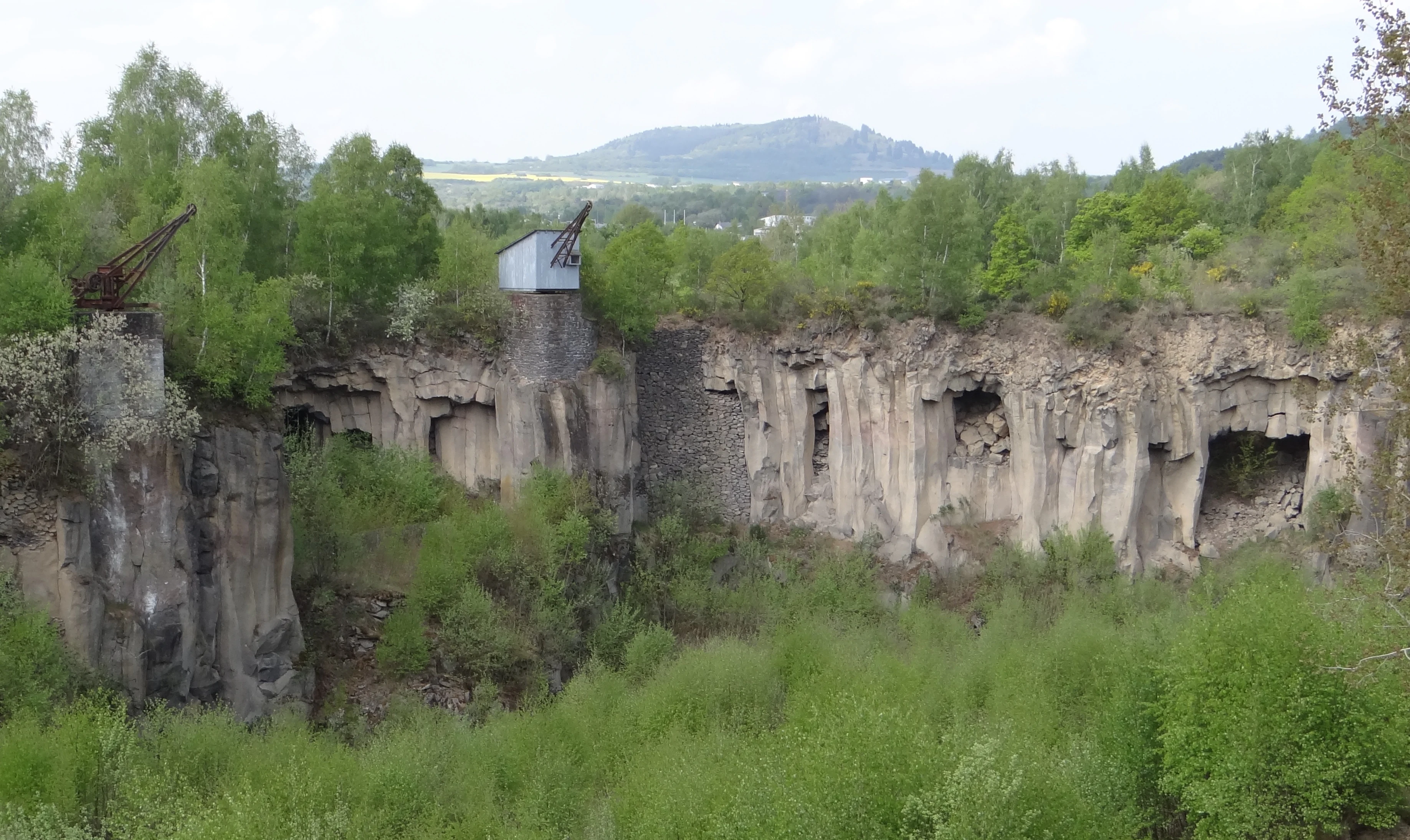 A wide view of a former basalt quarry featuring high rocky walls with indentations, rusted historic cranes, and lush green vegetation growing throughout the site.
