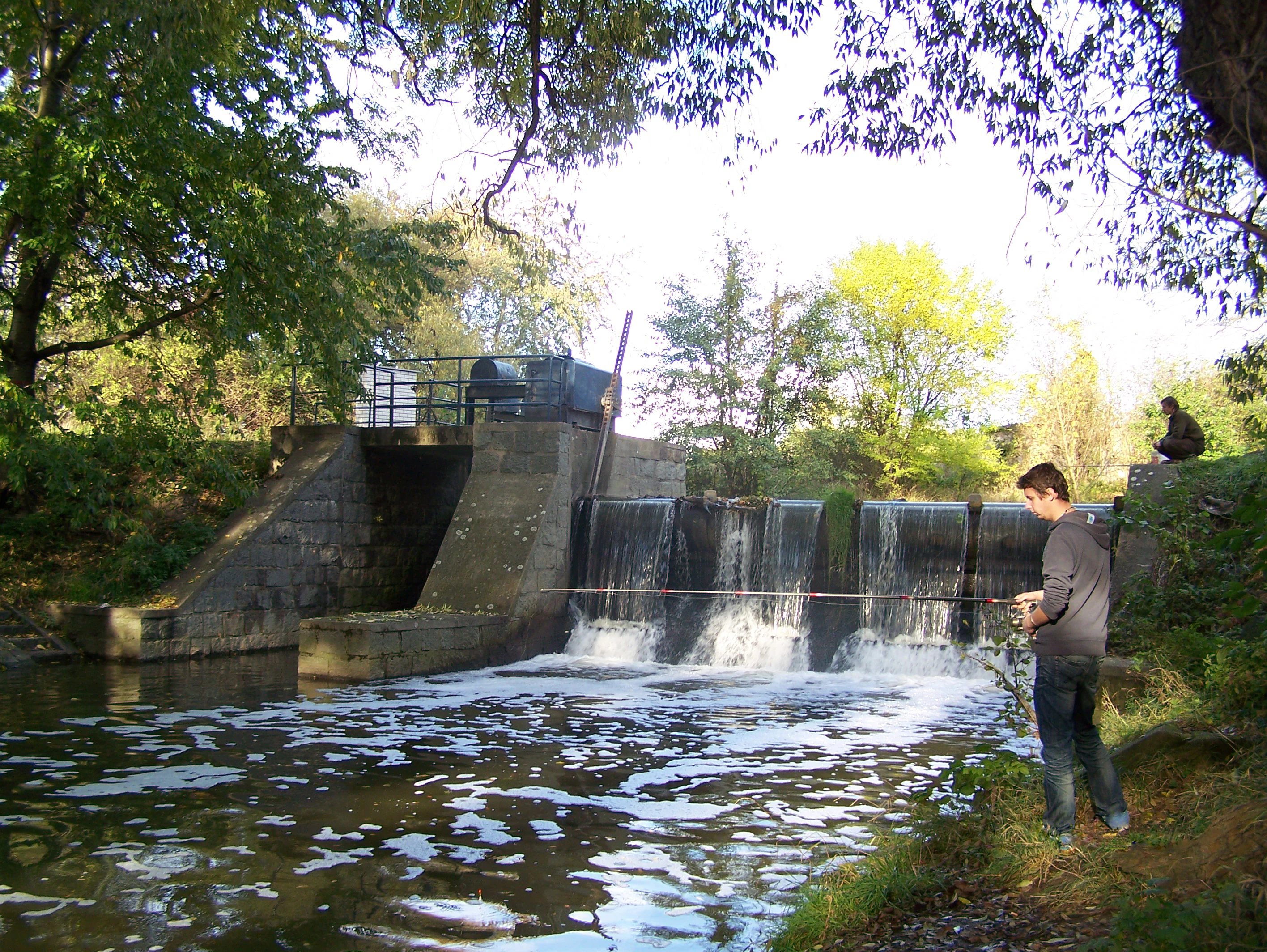 A man fishing with a rod on the bank of a small stream next to a stone and concrete weir, surrounded by trees.