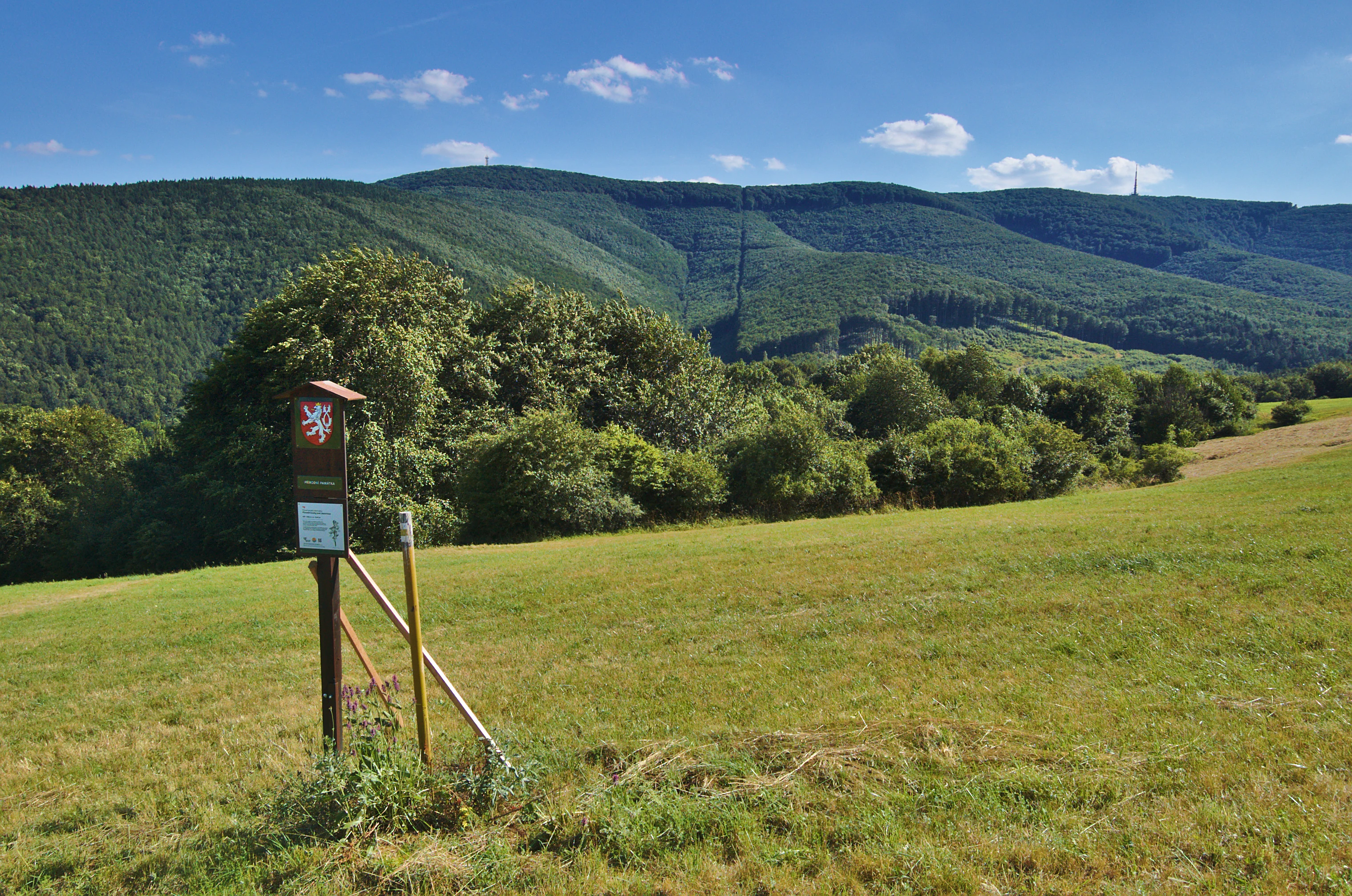 A nature reserve sign with a red shield emblem featuring a double-tailed lion sits on a wooden post in a grassy field, with forested hills in the background.