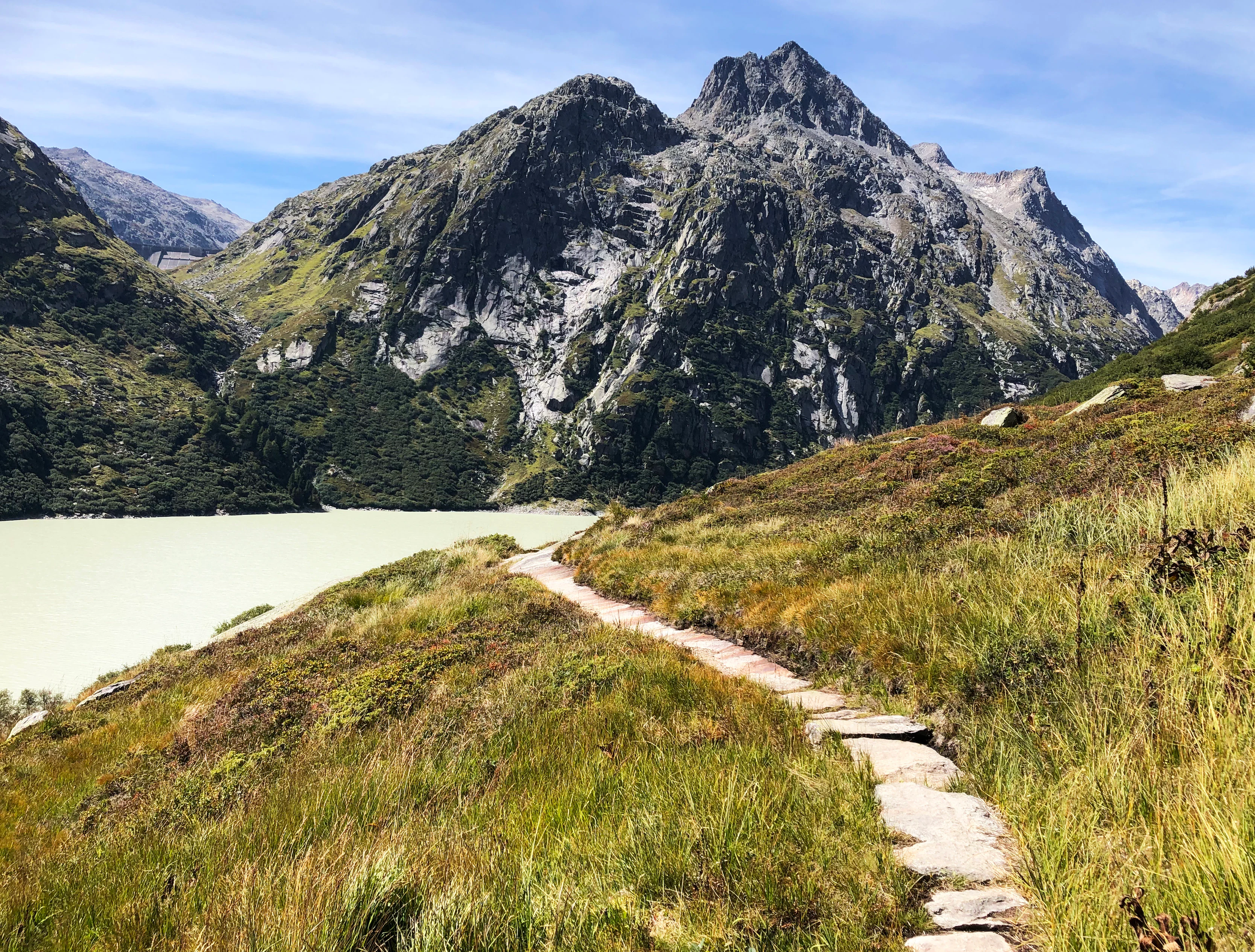 A narrow stone-paved trail curves through a grassy, alpine hillside toward a large, craggy mountain peak; a pale, turquoise-colored glacial lake is visible to the left.