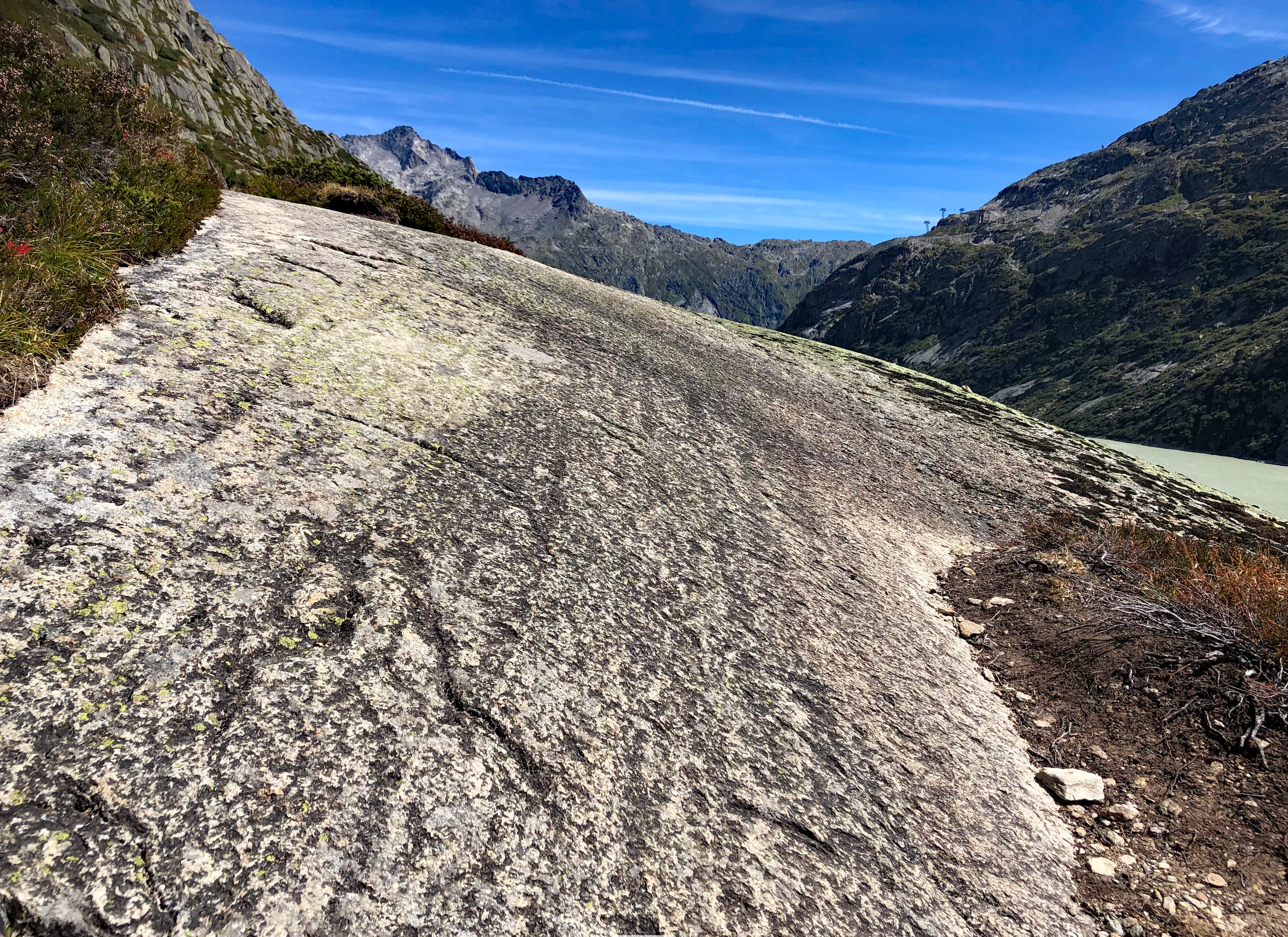 A wide angle shot of a large, smooth glacial-polished rock slab in the Swiss Alps, with mountainous terrain and a section of a turquoise lake visible in the background.