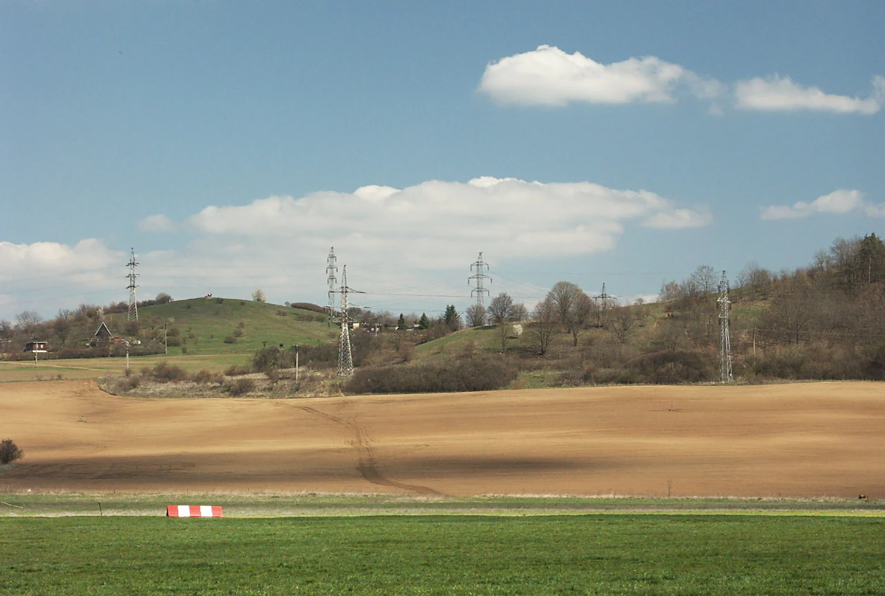 A wide, outdoor landscape shot featuring rolling hills with sparse trees and multiple electrical transmission towers in the distance, set behind a large field of tilled soil and a green grassy foreground.