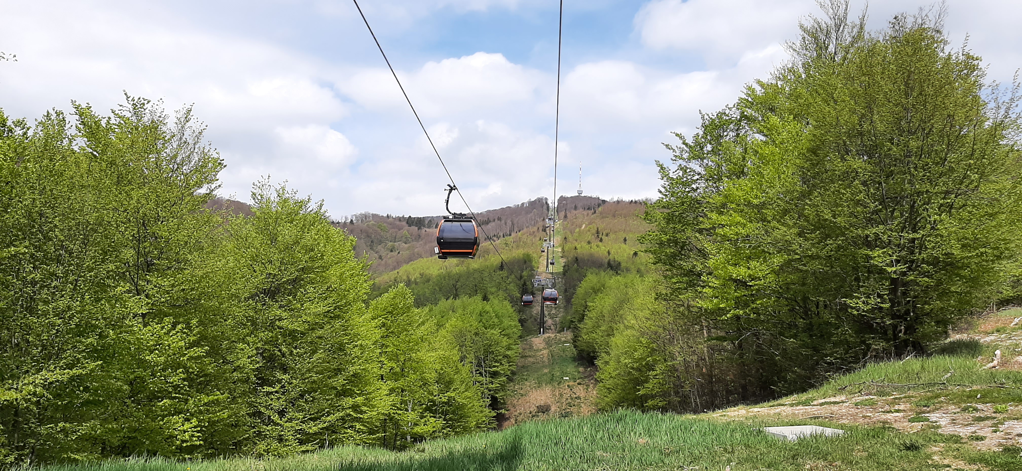 A cable car suspended between two towers, surrounded by green forest and rolling hills under a partly cloudy sky
