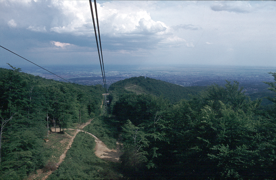 Cable car suspended above forested mountain slope with dirt path, overlooking cityscape under partly cloudy skies