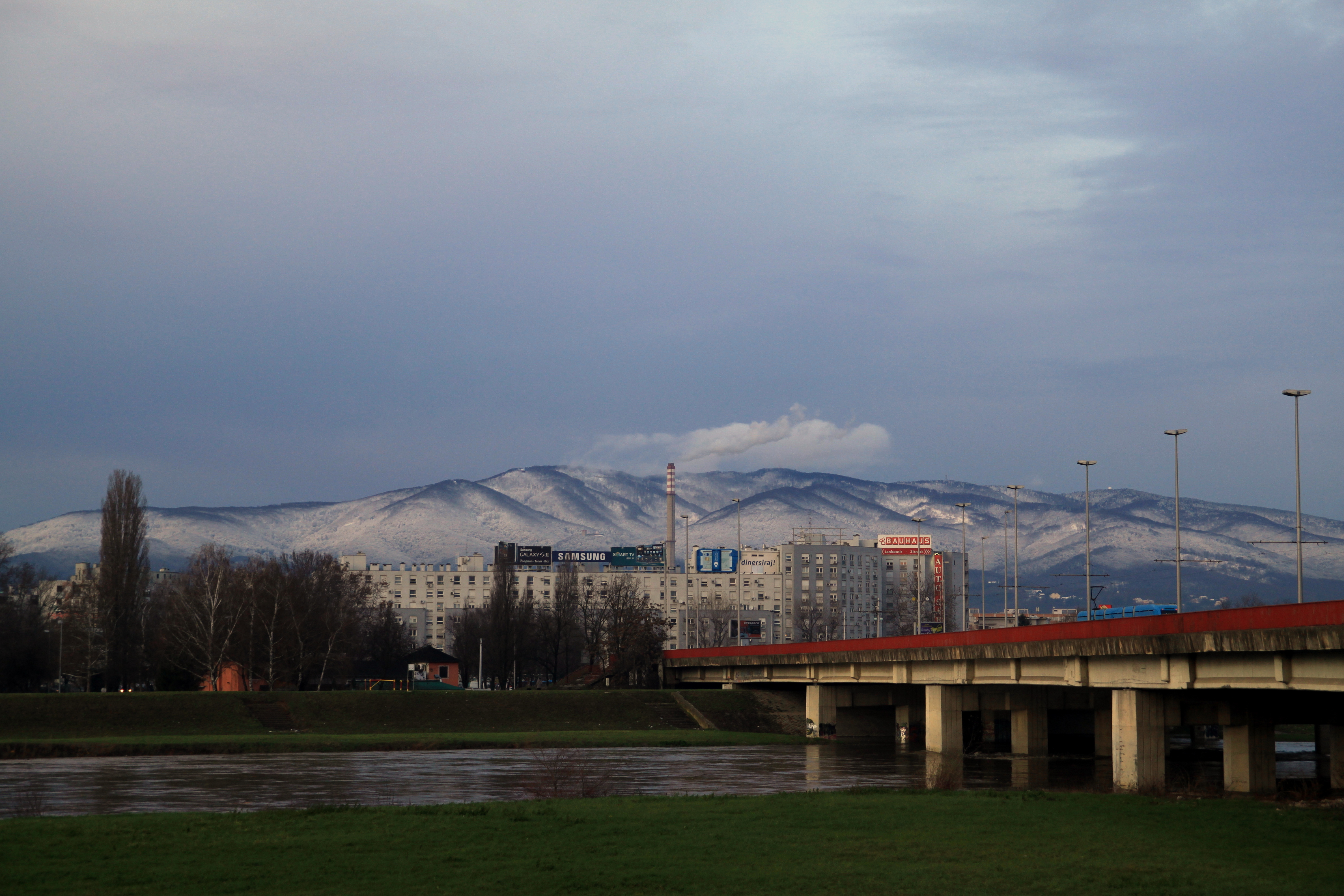 Snow-capped Medvednica mountain range visible from a riverbank with a bridge, buildings, and grassy foreground under an overcast sky