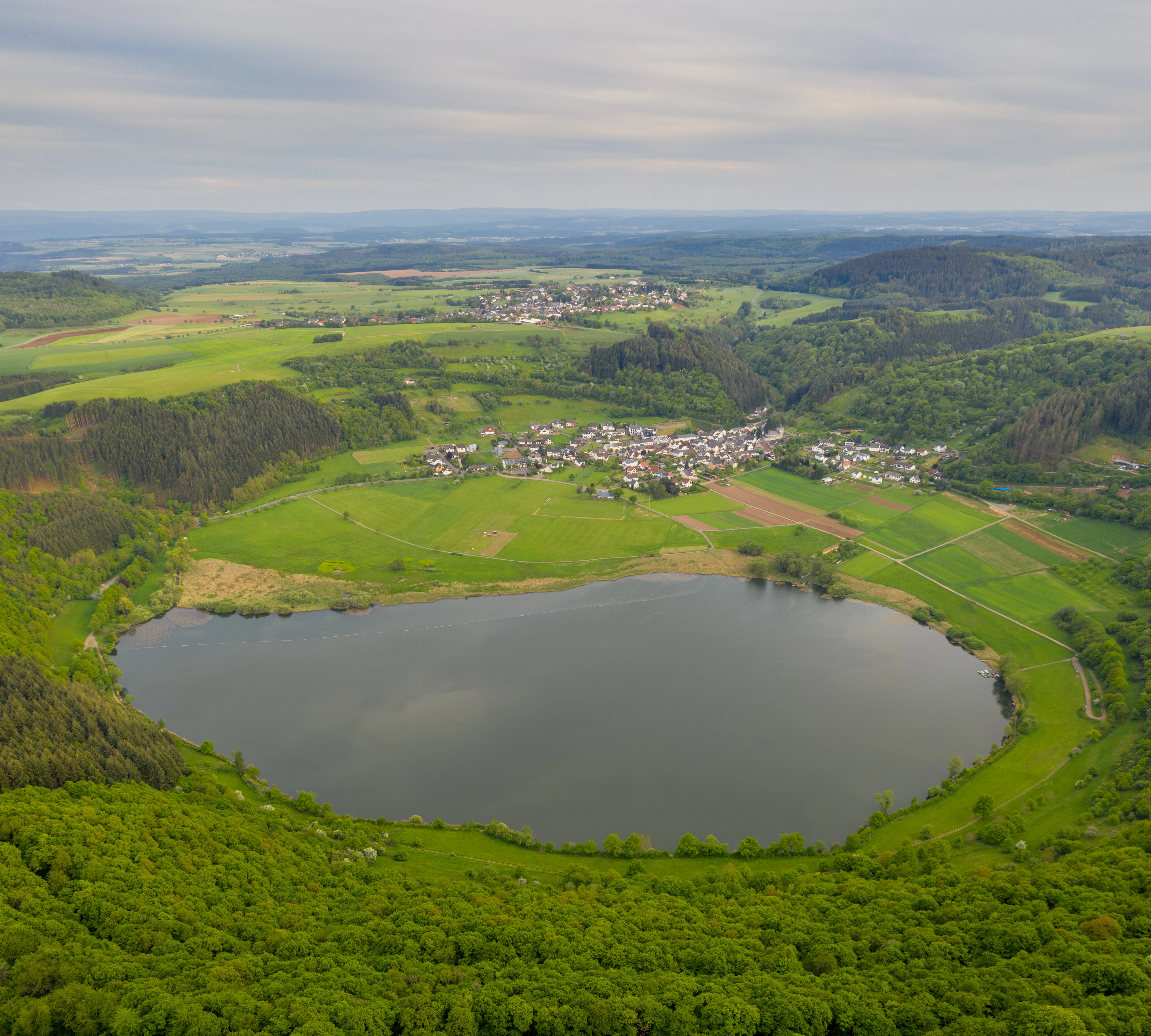 A wide aerial view of a circular volcanic crater lake surrounded by green forests, grassy fields, and the small village of Meerfeld.