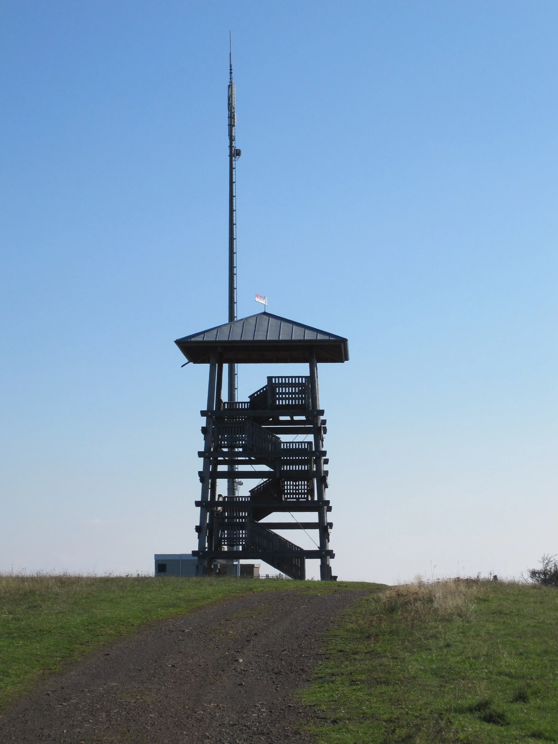 A wooden observation tower stands on a grassy hill under a clear blue sky, with a tall telecommunications mast rising behind it.