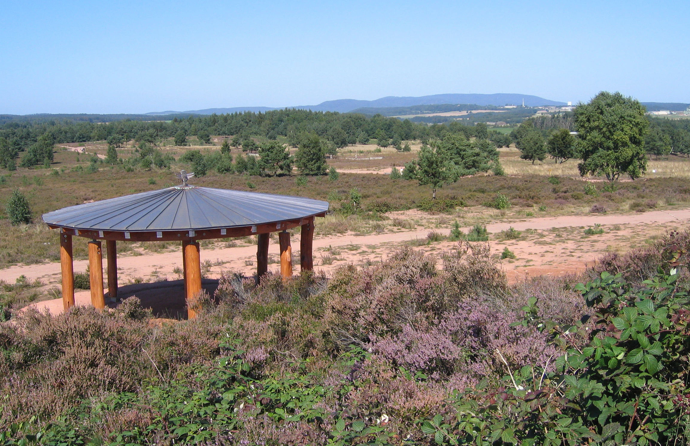 A circular wooden pavilion with a metal roof stands on a dirt path in a heathland area with purple heather, scattered trees, and distant hills under a clear blue sky.